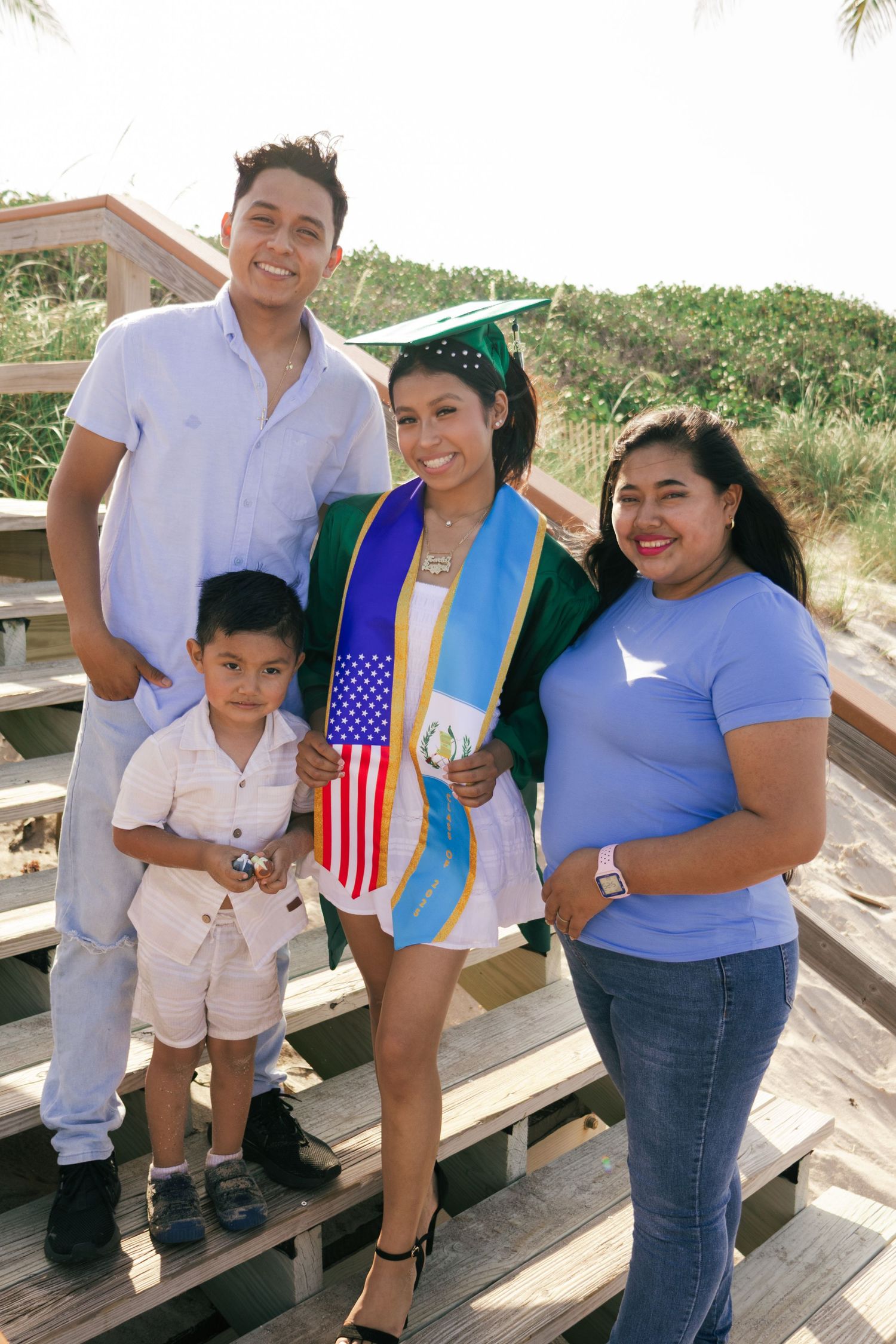 Family celebrates graduation outdoors, graduate in green cap and gown with US and Guatemalan flag sashes poses with loved ones.