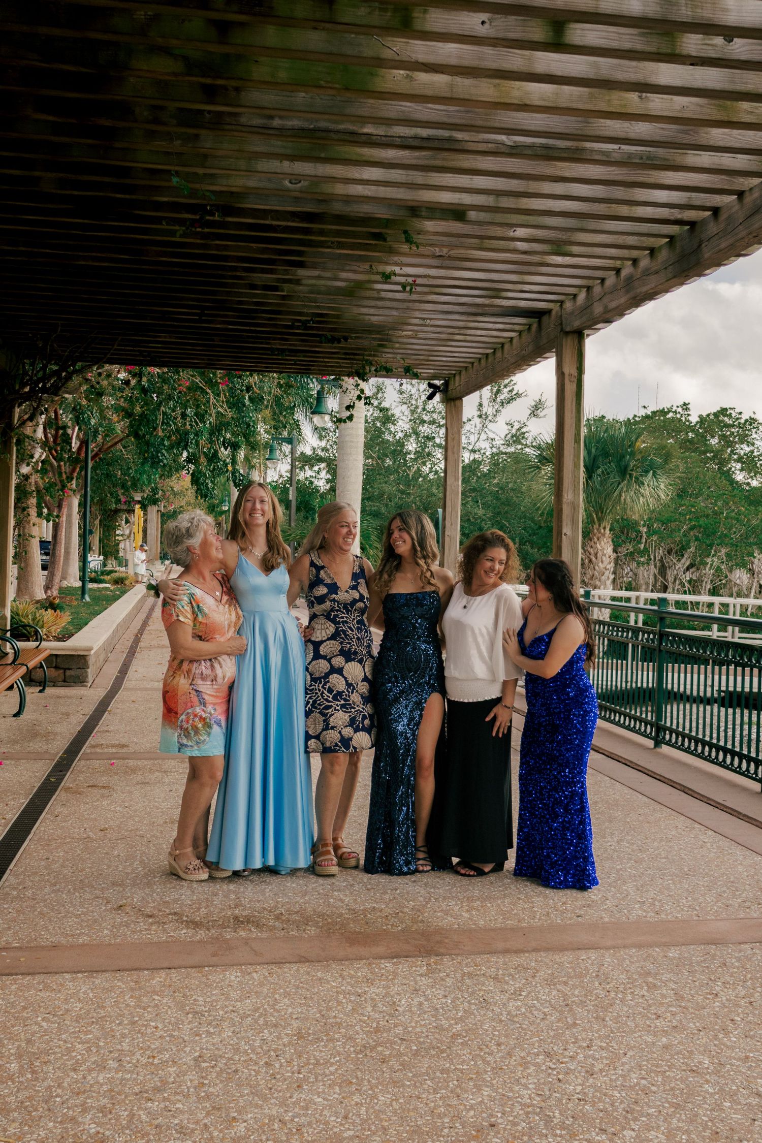 Five women in formal dresses pose together under a pergola walkway at an outdoor venue.