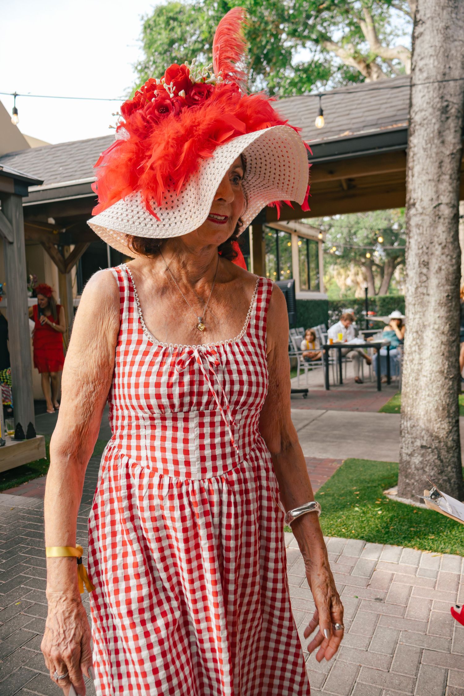 Elegant woman in red gingham dress and wide-brim hat with red feathers at an outdoor garden party.