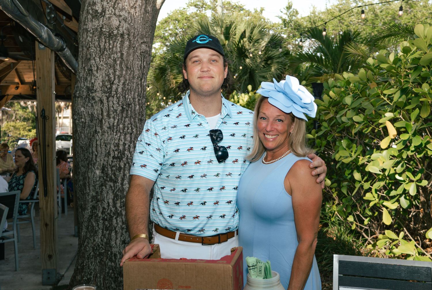 A smiling couple poses outdoors at a garden party, man in blue polo and woman in light blue dress with matching hat.