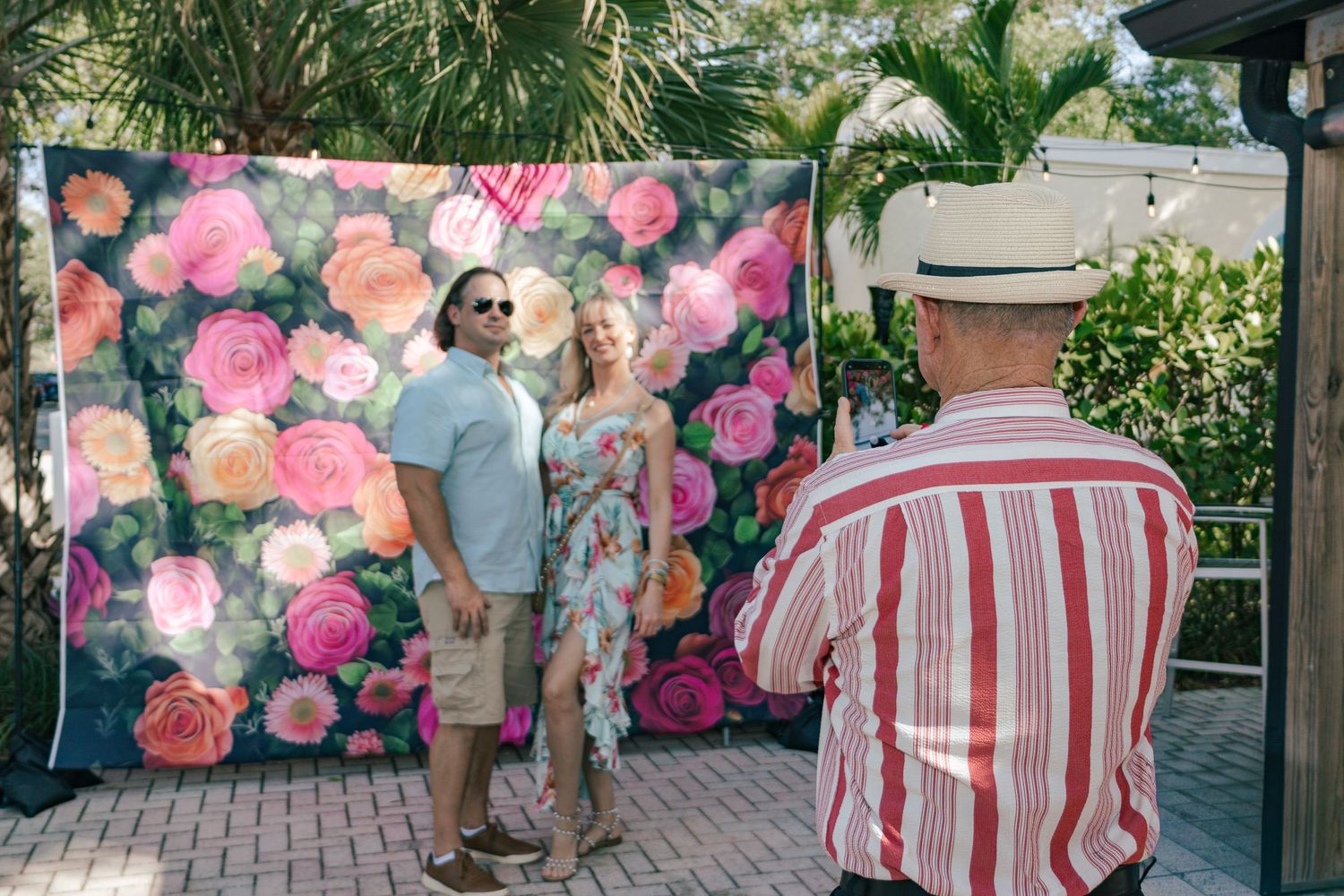 Couple posing in front of a floral backdrop while a photographer in a striped shirt captures the moment.