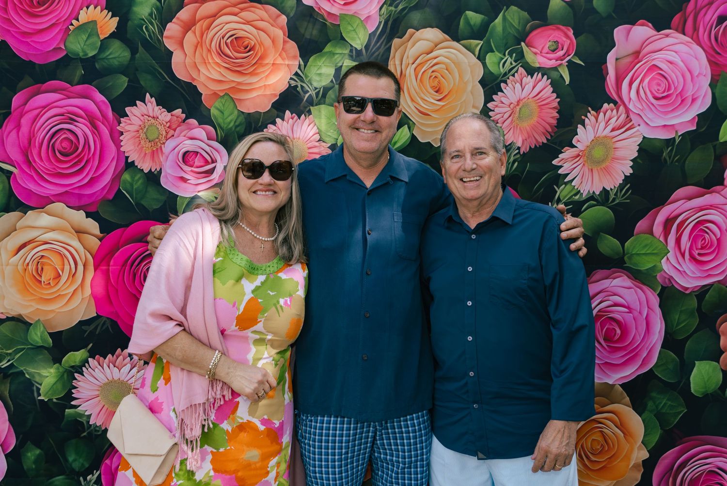 Three people smiling in front of a colorful floral backdrop with pink, orange, and red roses.