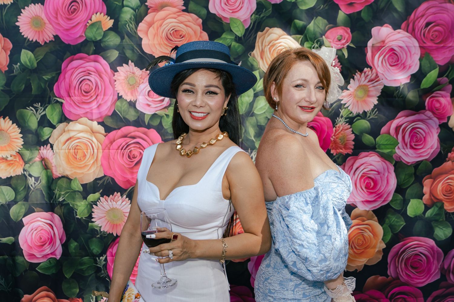 Two elegant women in hats and formal dresses posing back-to-back before a vibrant painted floral rose backdrop.