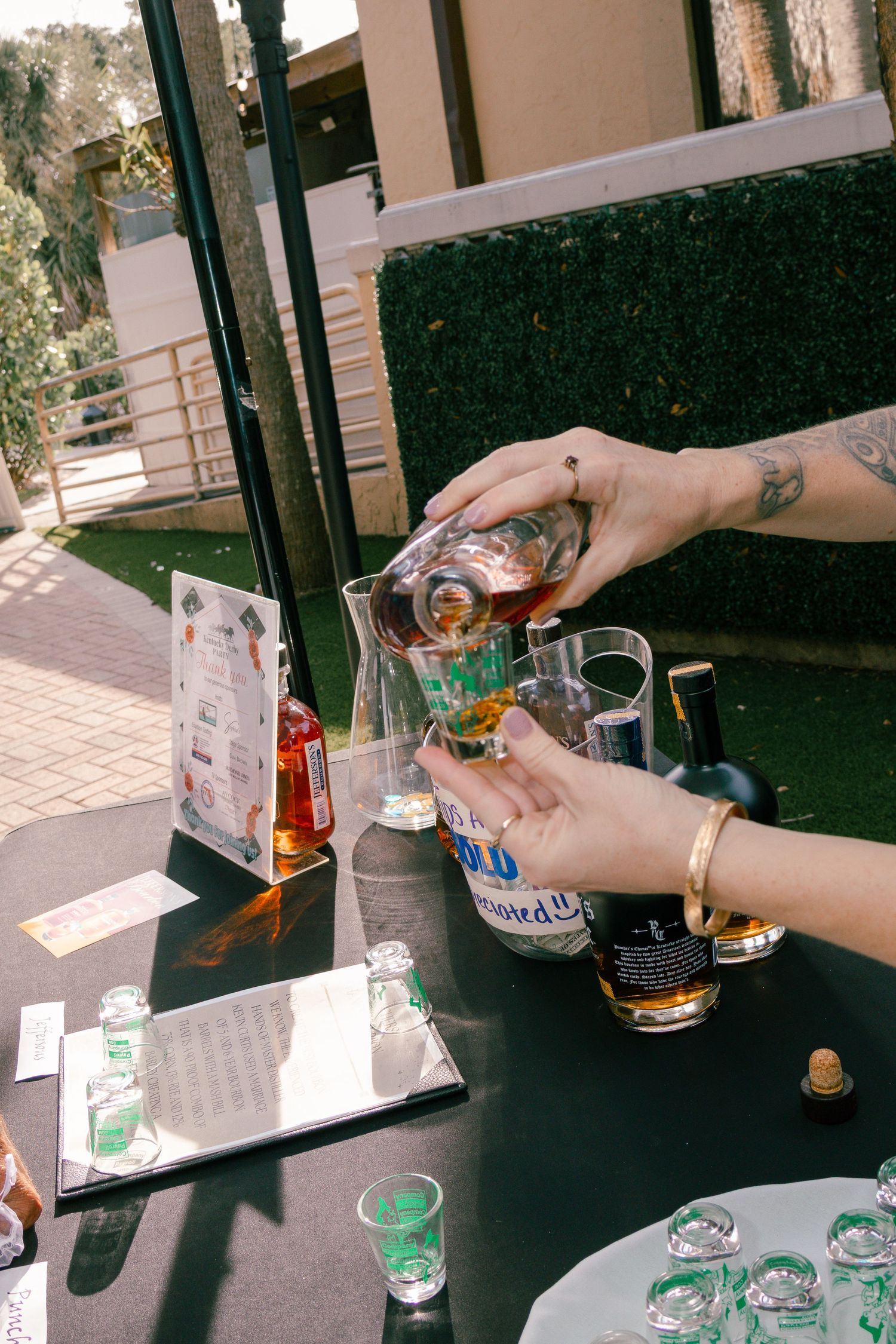 Hands pouring whiskey into a glass at an outdoor Derby party bar setup.