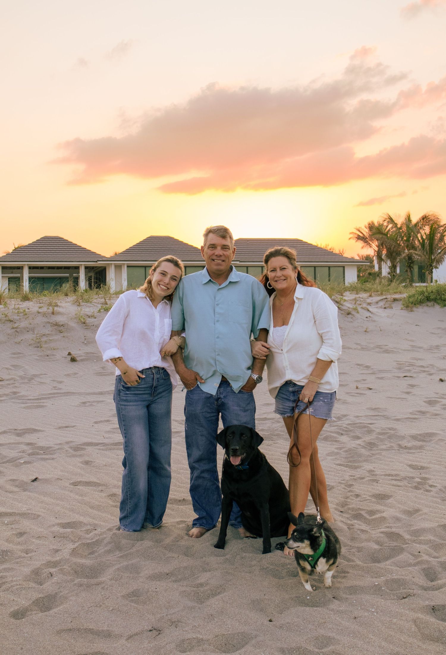 Family of three posing with two dogs on a sandy beach during a vibrant sunset.