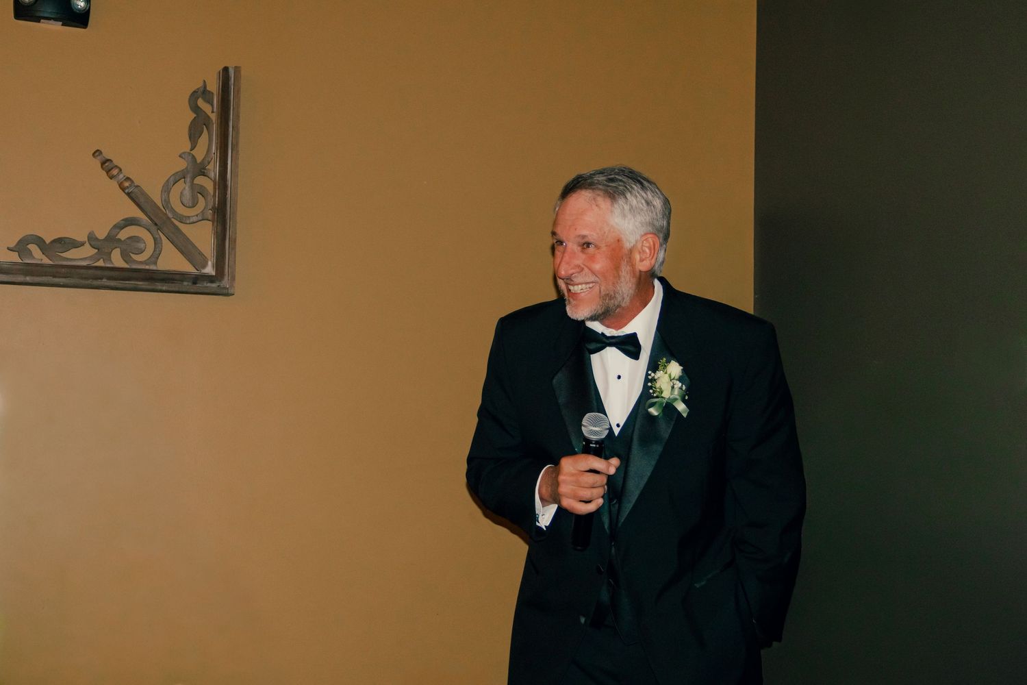 Elegant older gentleman in black tuxedo with boutonniere delivers a wedding speech while holding a microphone.