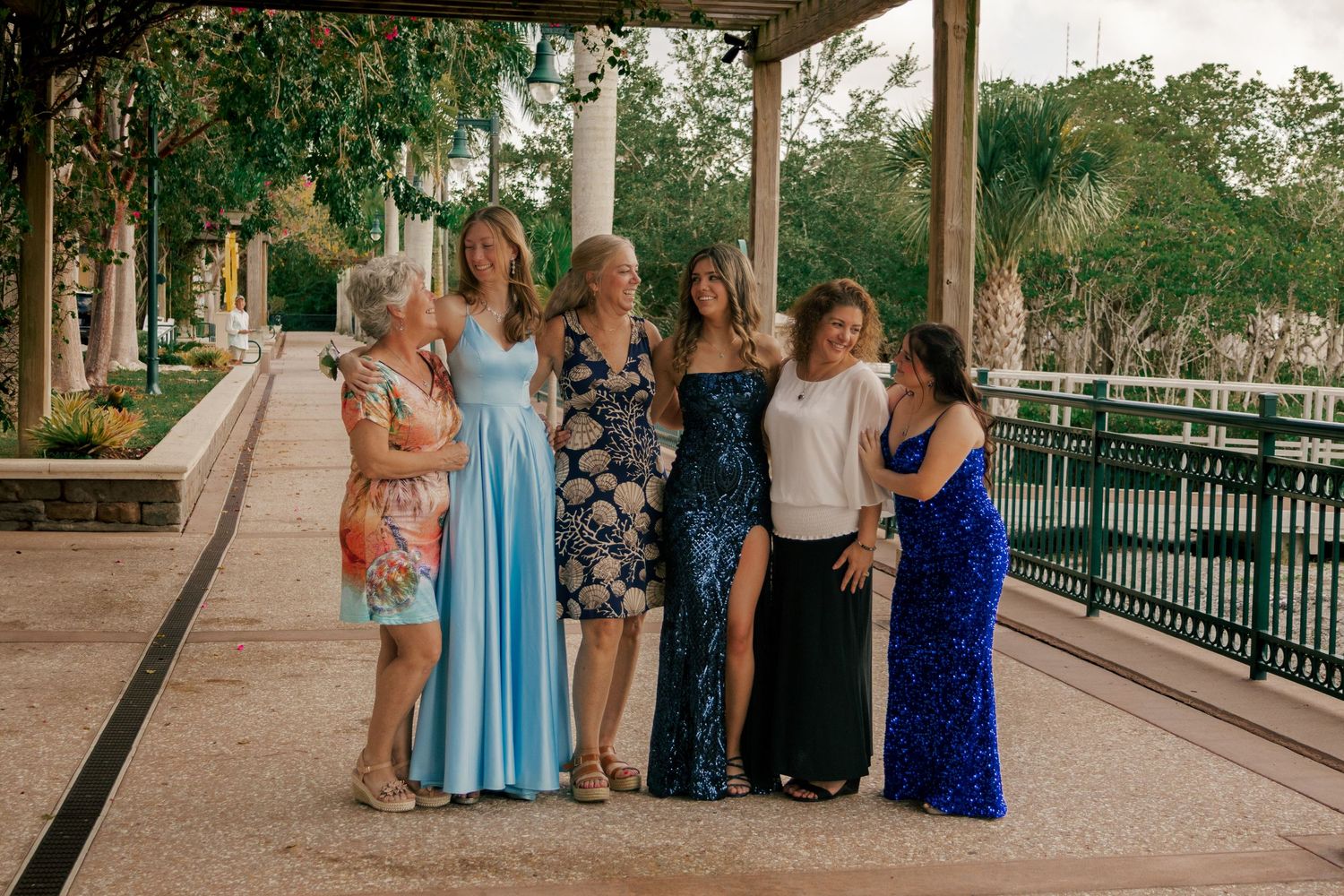 Six women of different ages smile and chat together under a tree-lined pergola walkway.