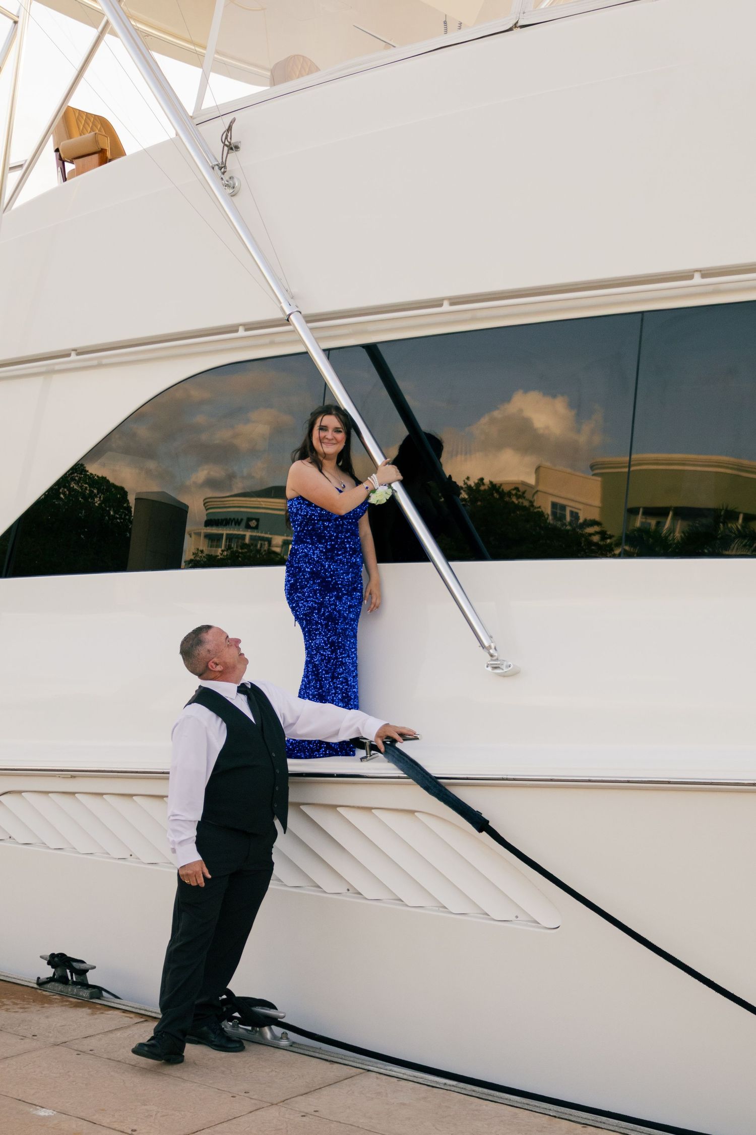 Man in black vest standing below woman in royal blue sequin gown posing on white yacht gangway.