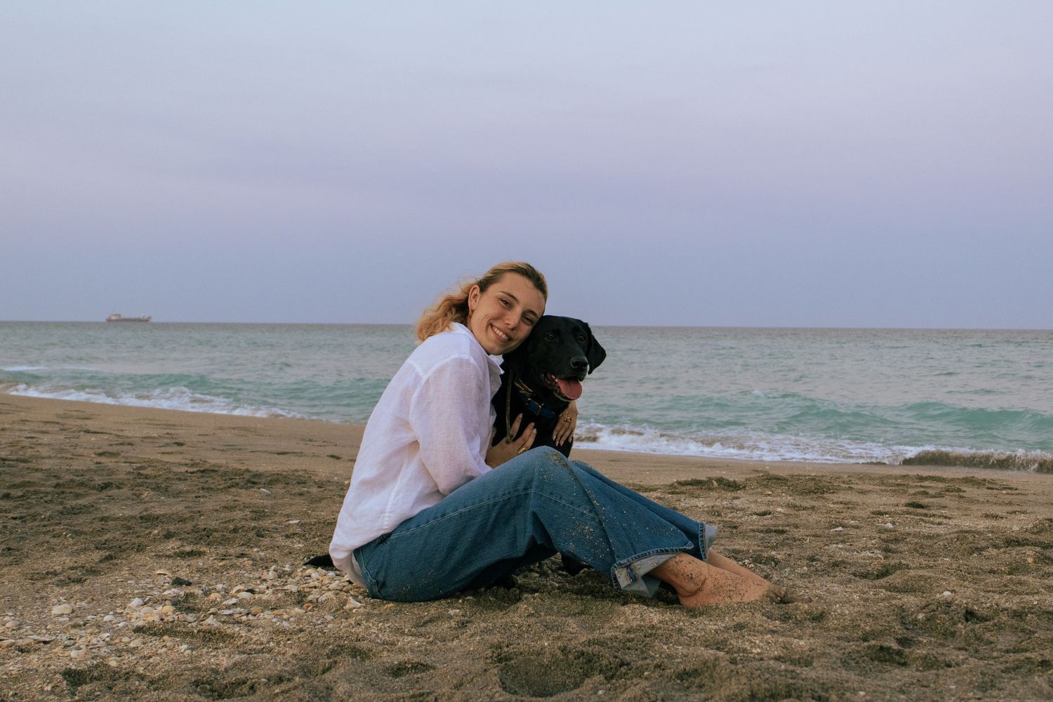 Young woman sitting on beach sand hugging a black dog with ocean in background at dusk.