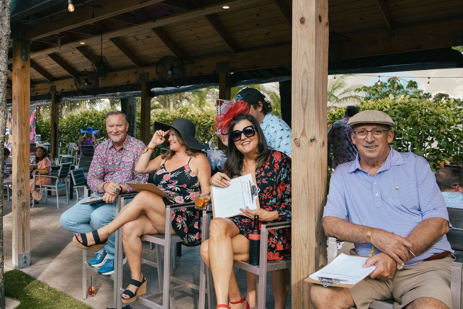 Group of guests in decorative hats relax under a wooden pergola at a Kentucky Derby watch party.