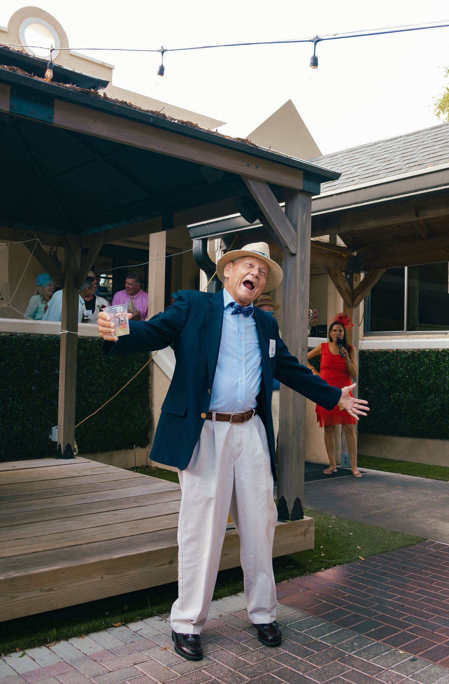 Elderly man in navy blazer, straw hat, and white pants gestures expressively outdoors at a festive gathering.