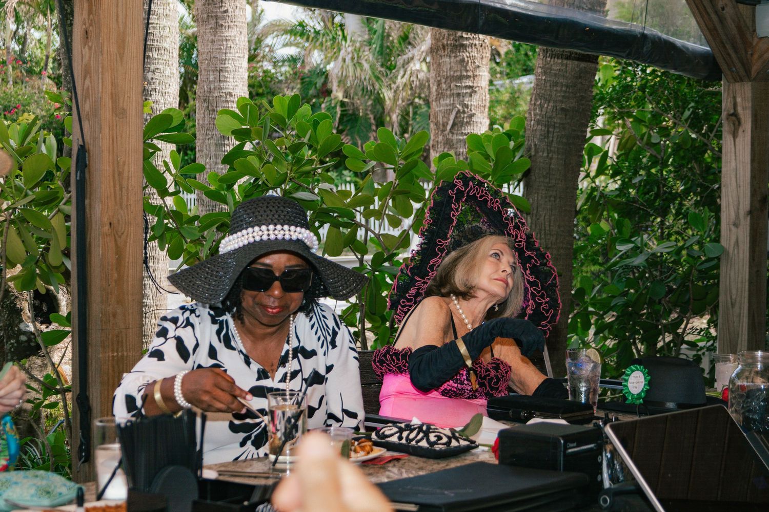 Two women in colorful hats and sunglasses sit together under a tropical cabana.
