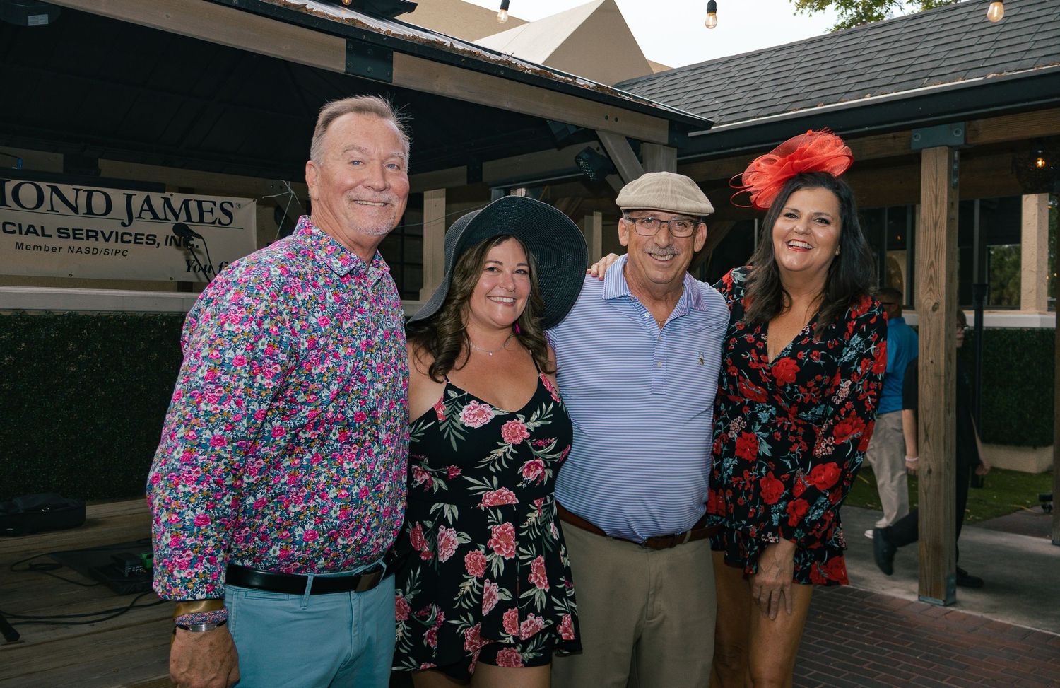 Two couples pose together outdoors wearing colorful floral outfits and festive hats.