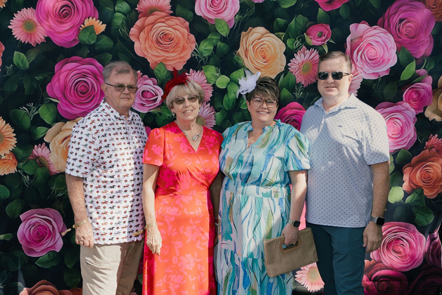 Four adults posing and smiling in front of a colorful floral backdrop with pink and orange roses.