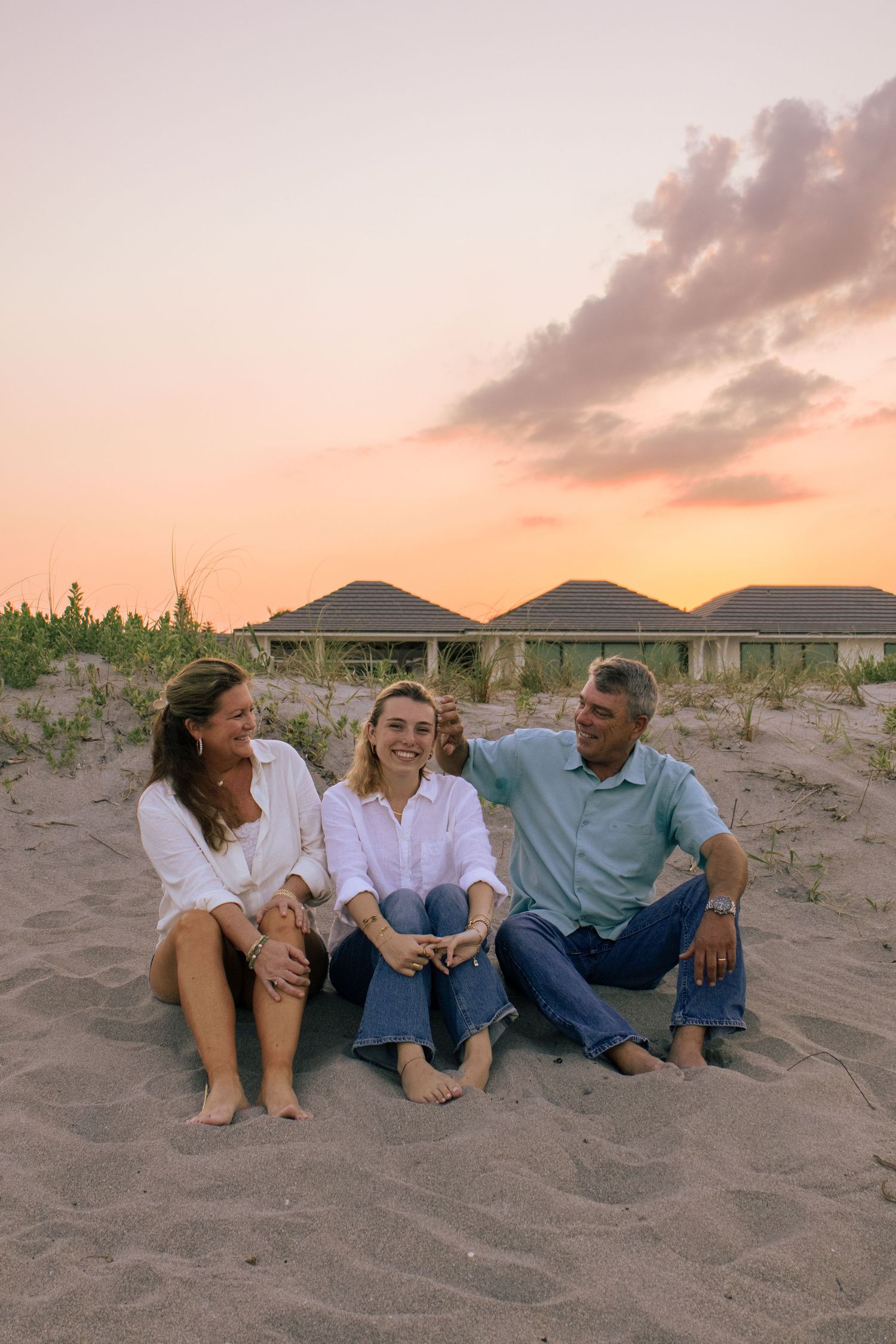 Family of three sitting barefoot on beach sand at golden sunset, laughing together near seaside dune cottages.