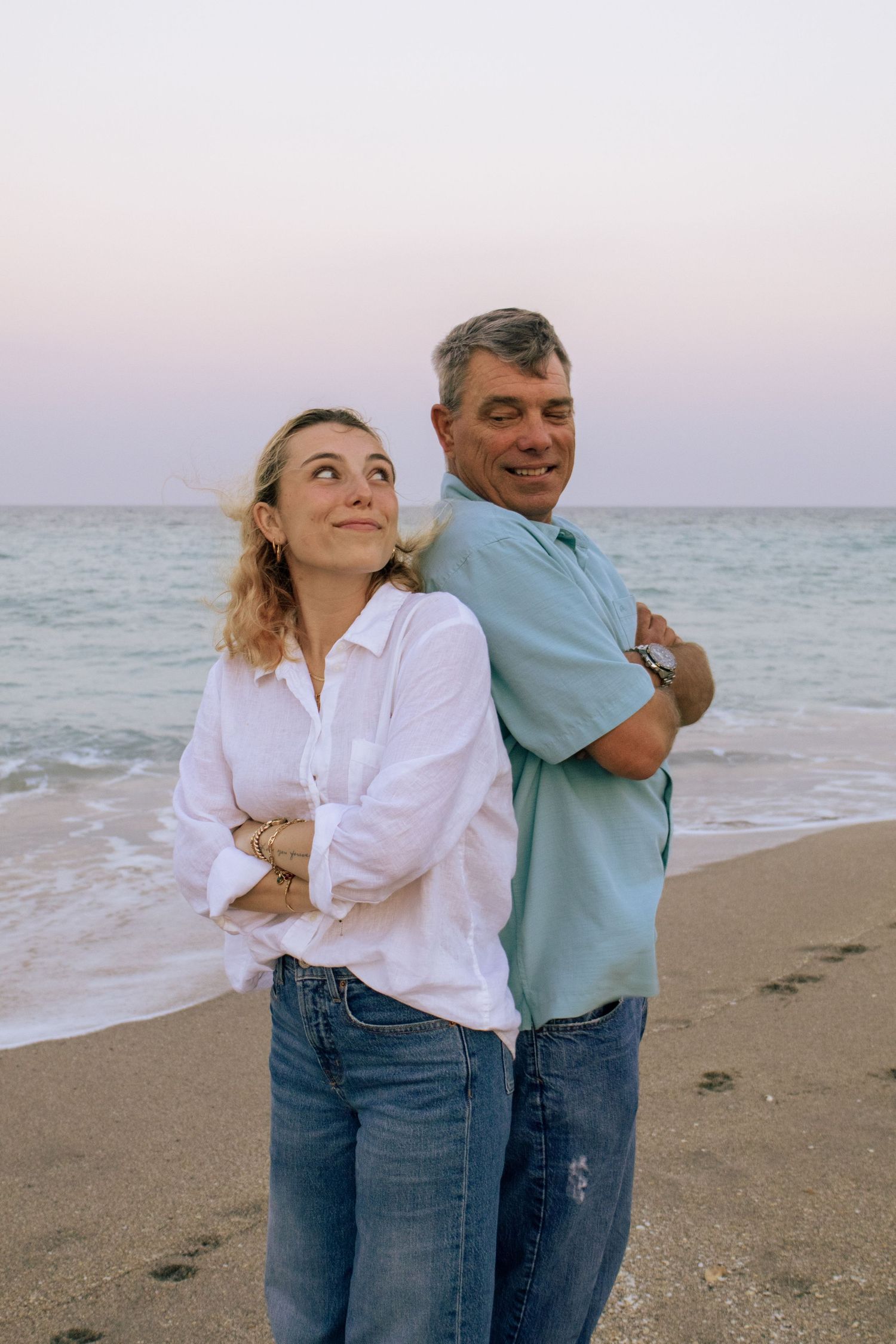 Young woman and older man standing back-to-back with arms crossed on sandy beach at dusk.