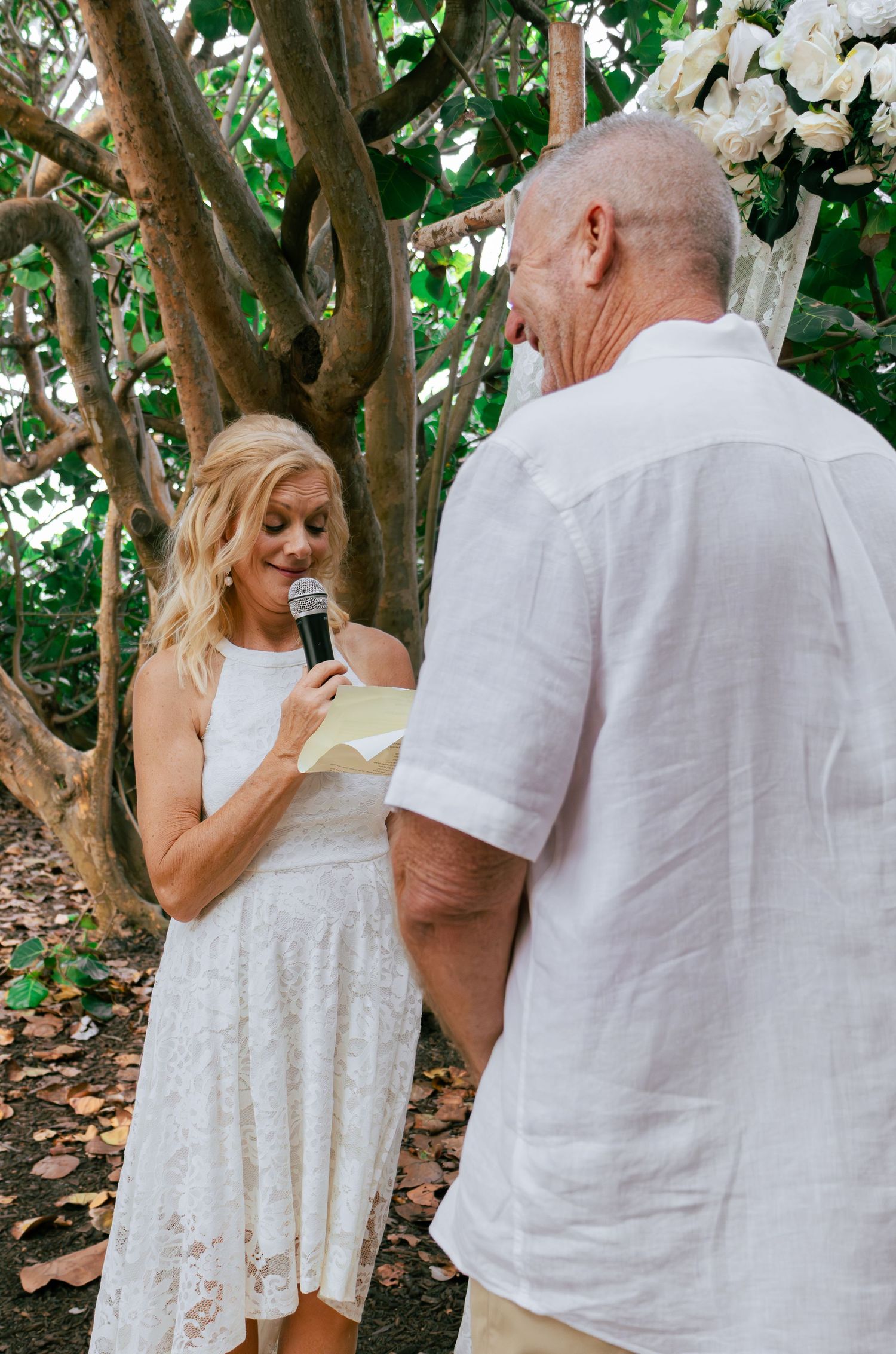 Blonde bride reading wedding vows into a microphone while facing her groom at an outdoor tropical ceremony.