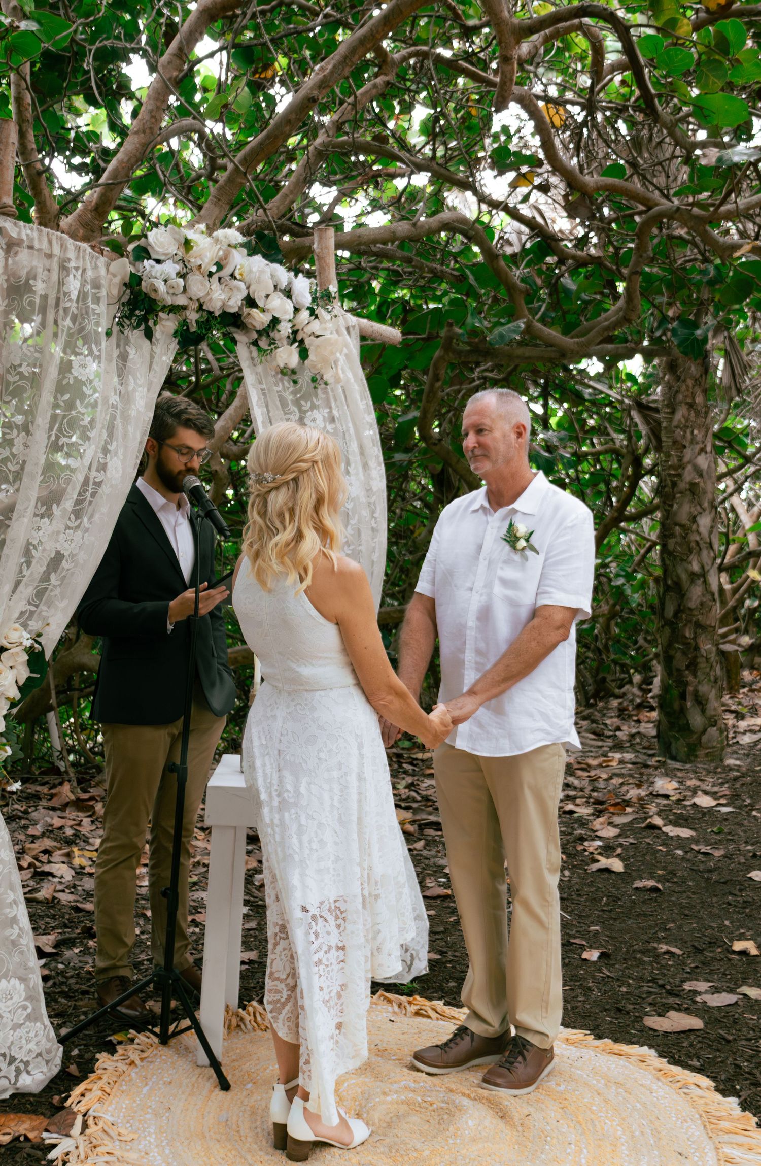 Bride and groom exchange vows under a floral arch with officiant in lush tropical garden setting.