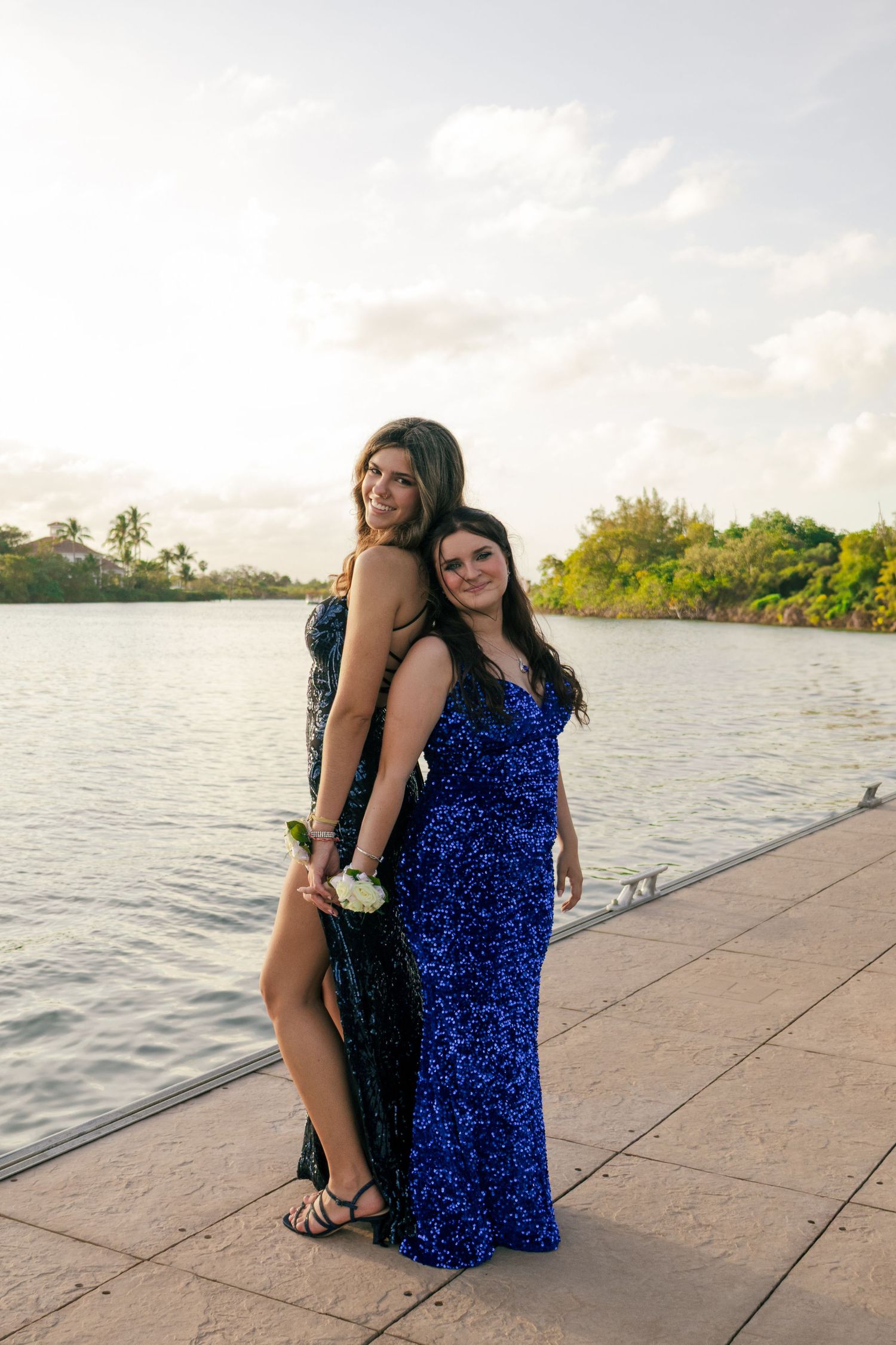 Two teenage girls in blue sequin prom dresses pose together on waterfront dock at sunset.
