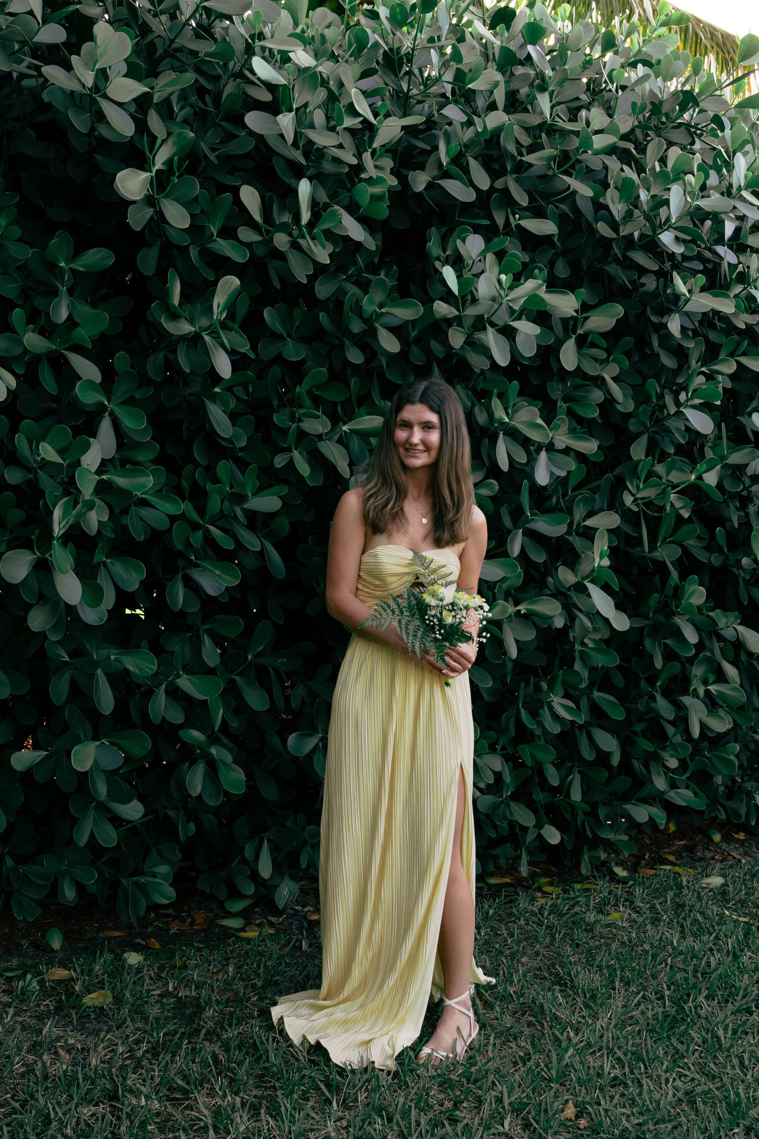 Young woman in a flowing yellow strapless gown holding a bouquet against a green hedge.