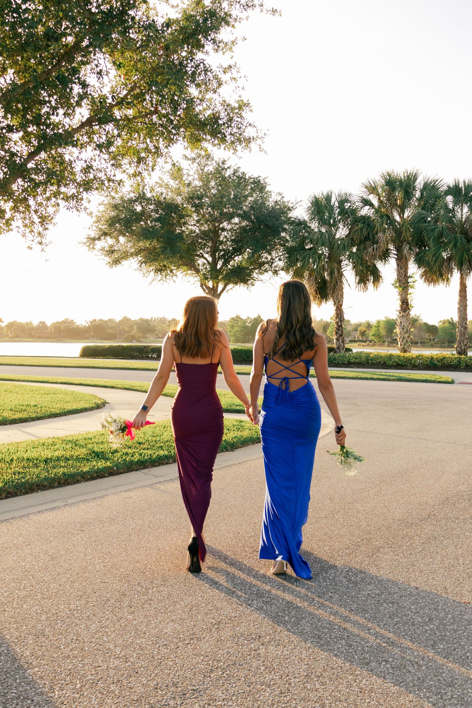 Two women in purple and blue formal gowns walk away holding flowers at golden sunset.