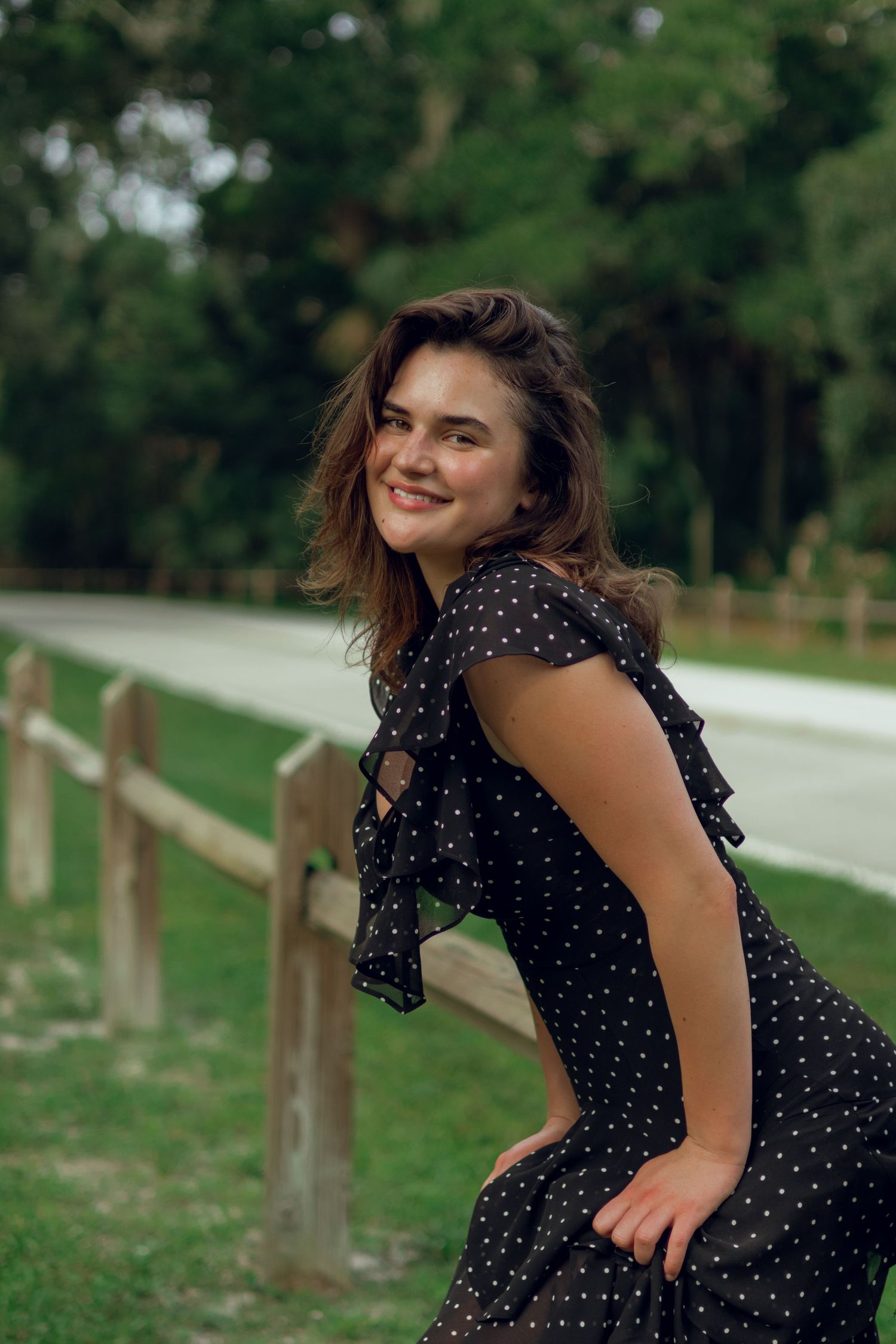 Smiling young woman in black polka dot dress leaning on wooden fence along a tree-lined road.