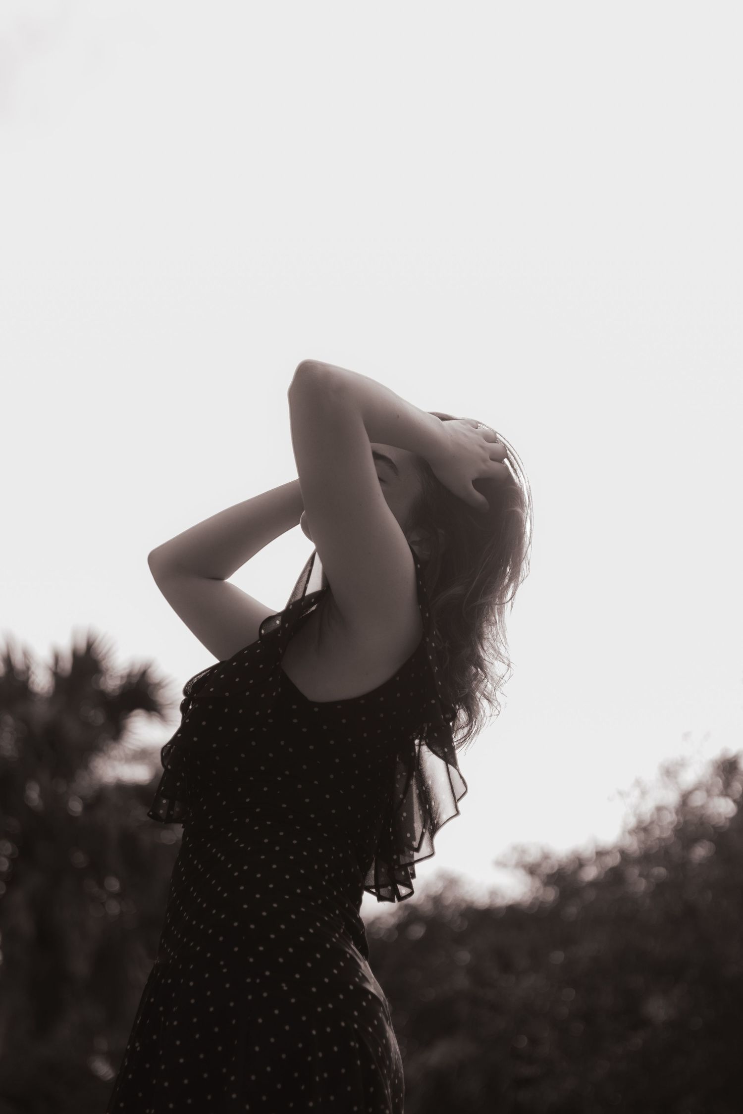 Silhouette of woman in black polka dot dress with hands in hair against bright sky, black and white.