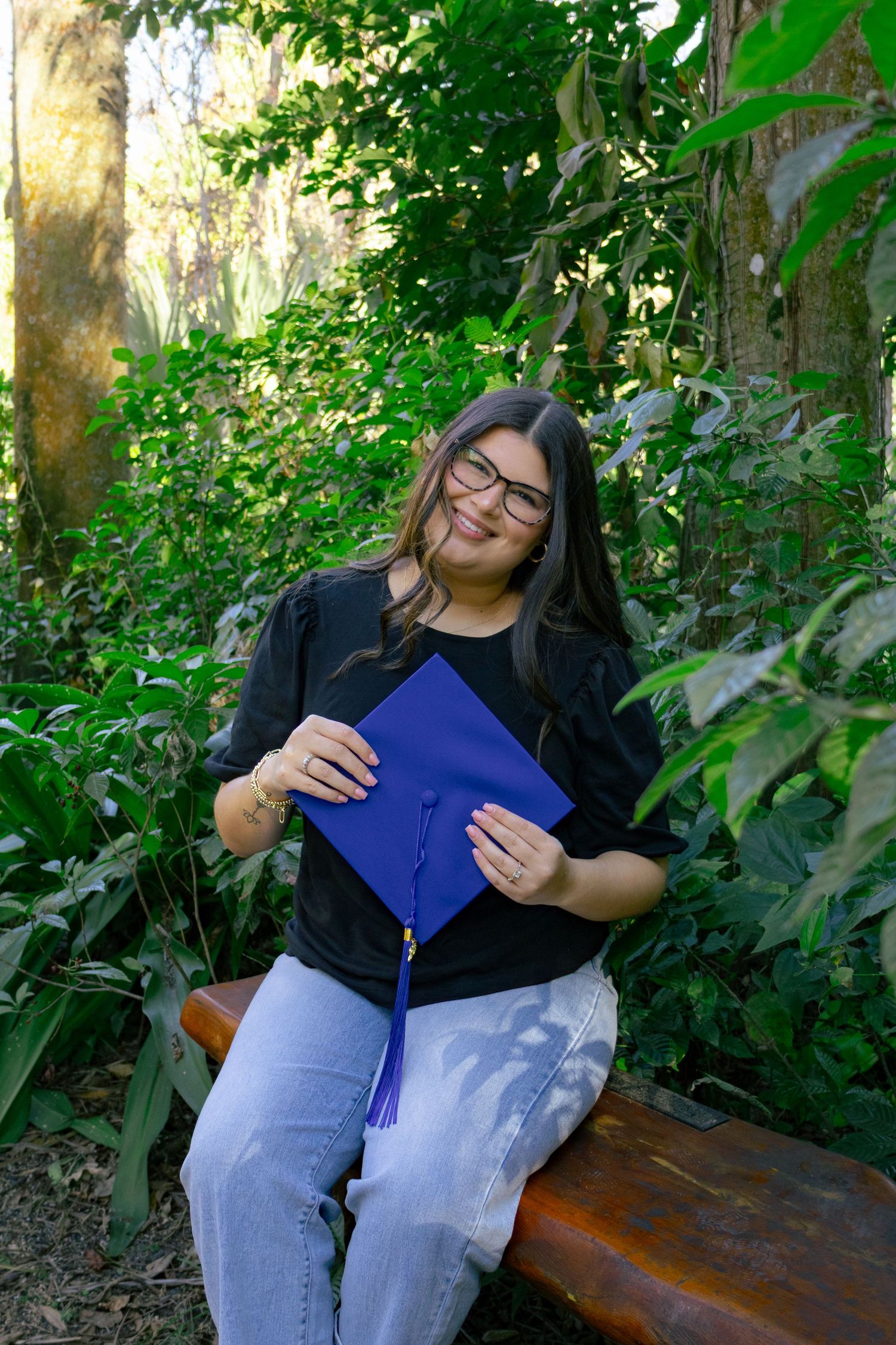 Smiling woman with glasses holds blue graduation cap while sitting on bench in lush garden.