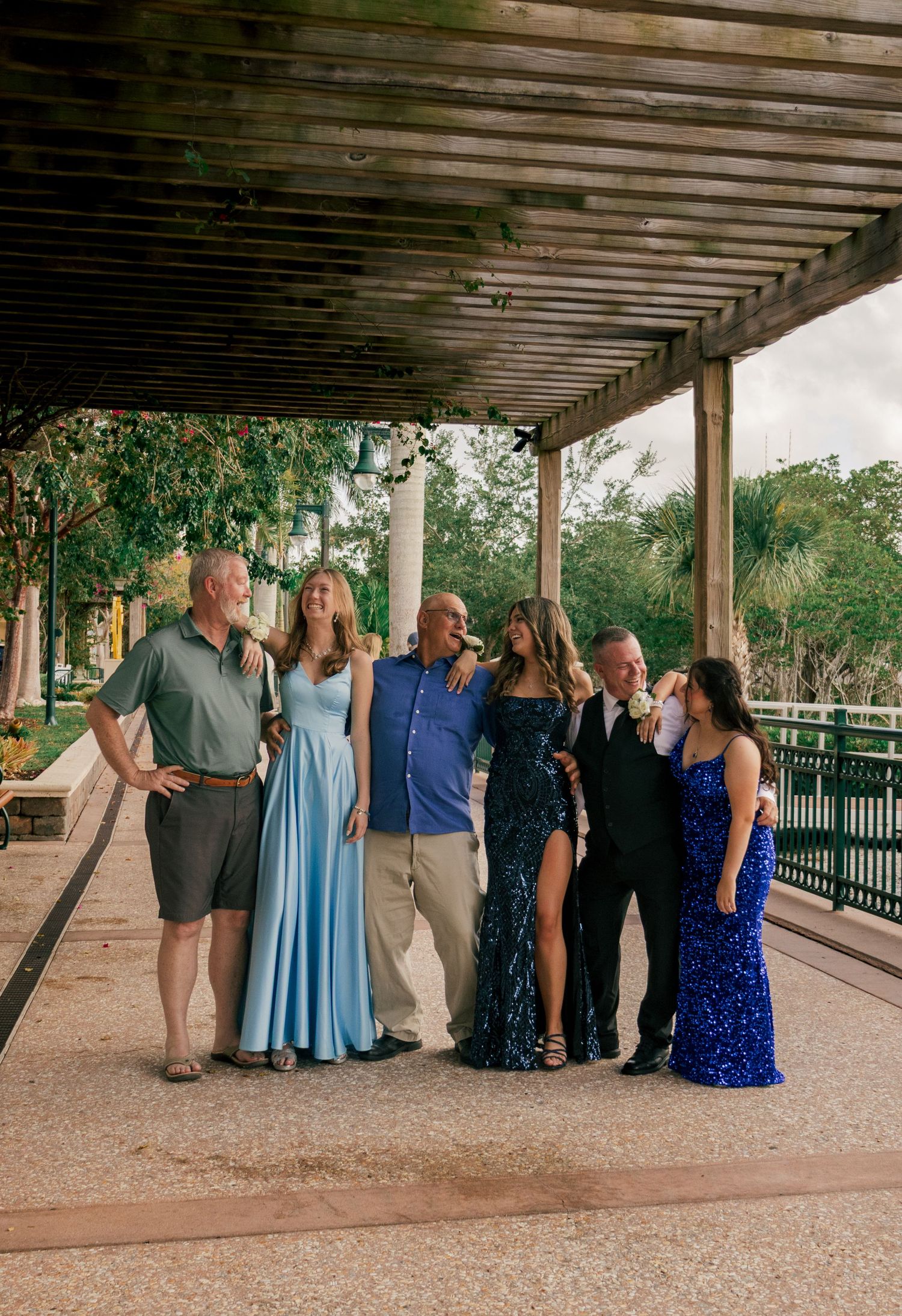 Mixed group of adults and teens in formal wear laugh together under a shaded pergola walkway.