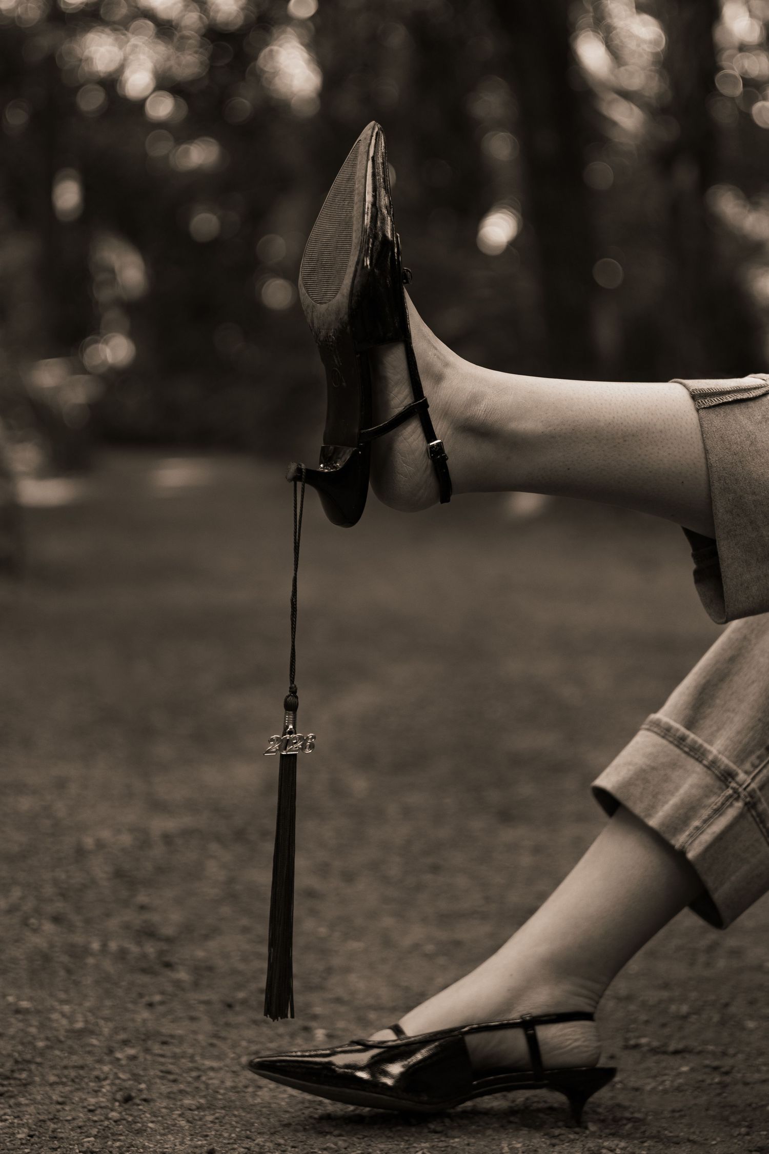 Black and white photo of a womans legs in heels with a dangling tassel keychain accessory on a forest path.