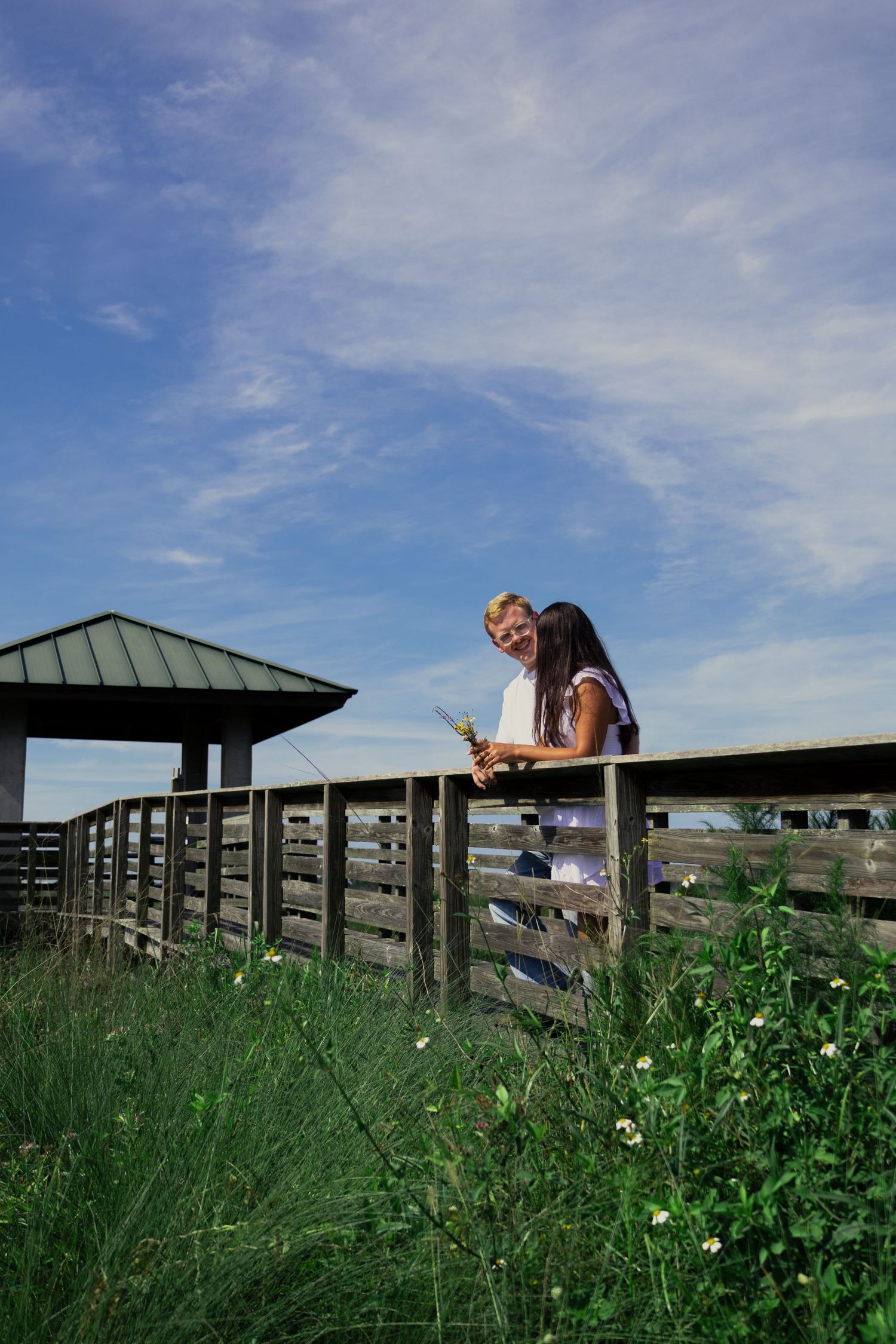 Romantic couple leaning on wooden fence railing on boardwalk surrounded by green meadow and blue sky.