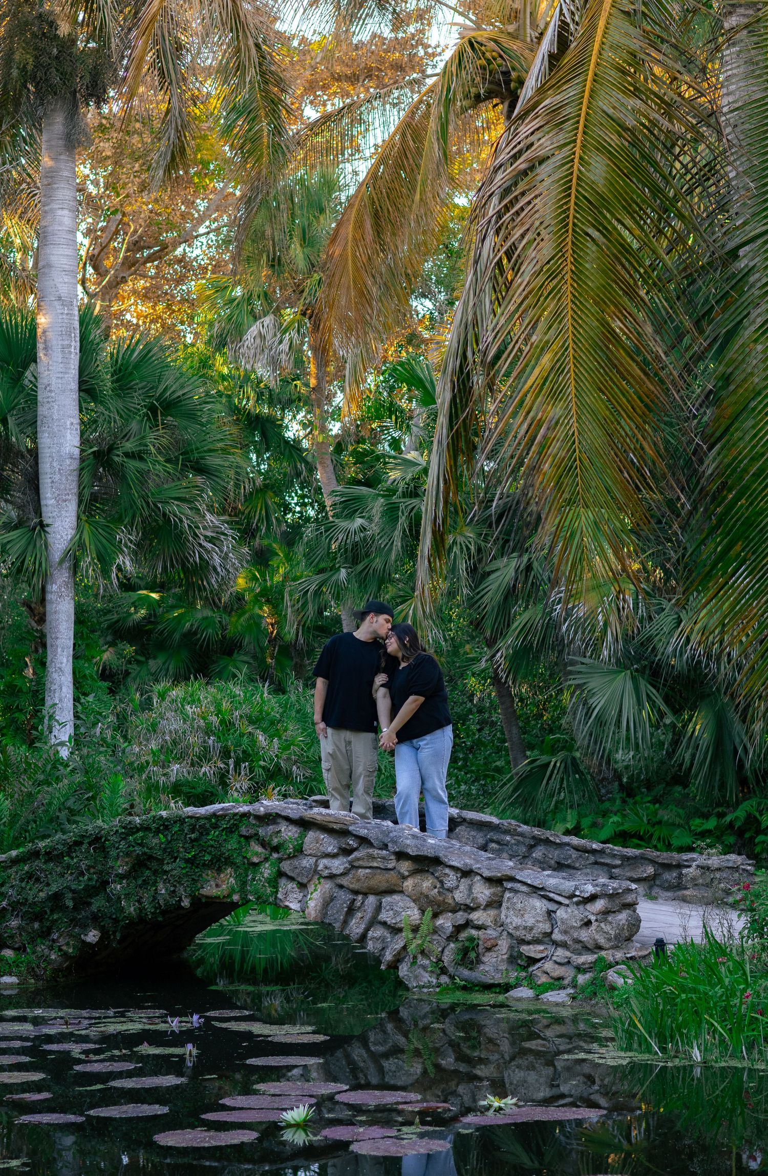 A couple holding hands and sharing a romantic moment on a rustic stone bridge surrounded by lush tropical foliage.