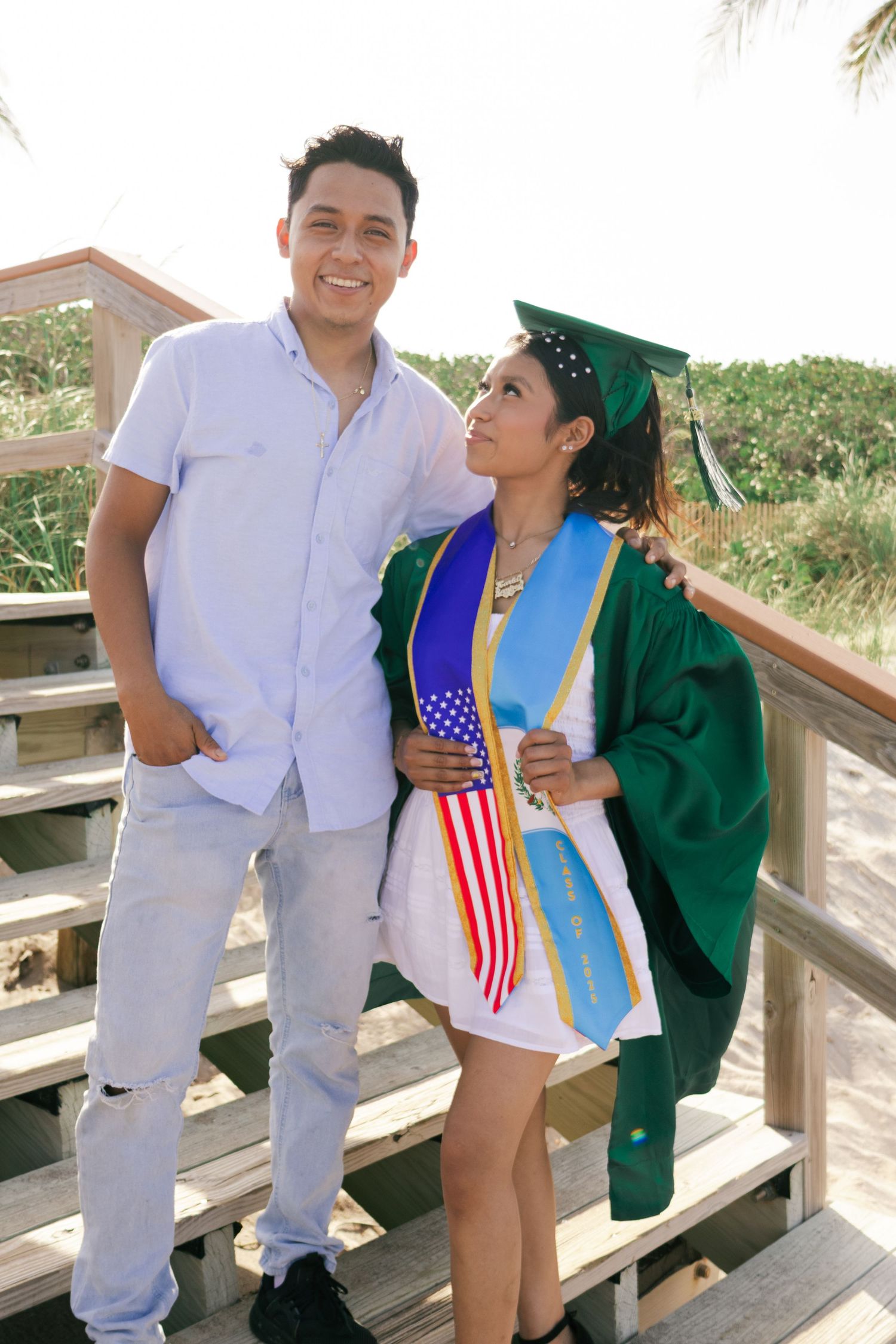 Young man and female graduate in green cap and gown pose together smiling on a sunny beach boardwalk.