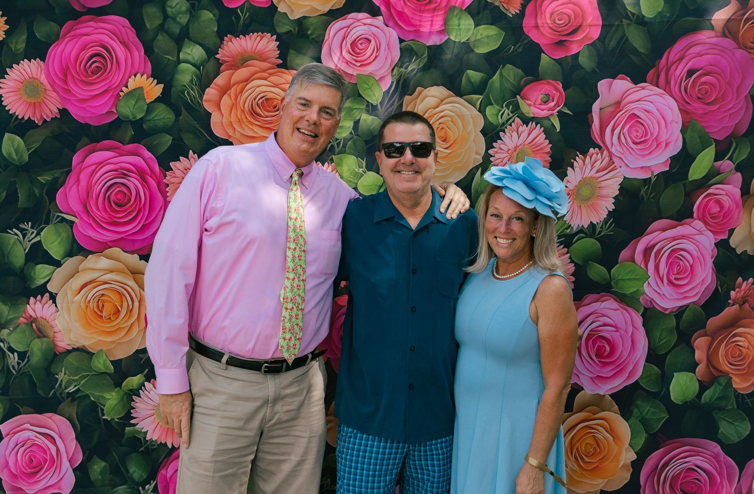 Three people smiling together in front of a colorful rose floral backdrop at a Derby party.