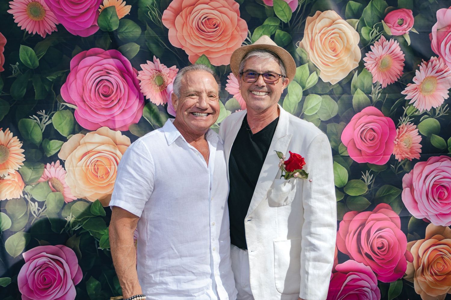 Two men smiling together in front of a colorful rose floral backdrop at a Derby celebration.