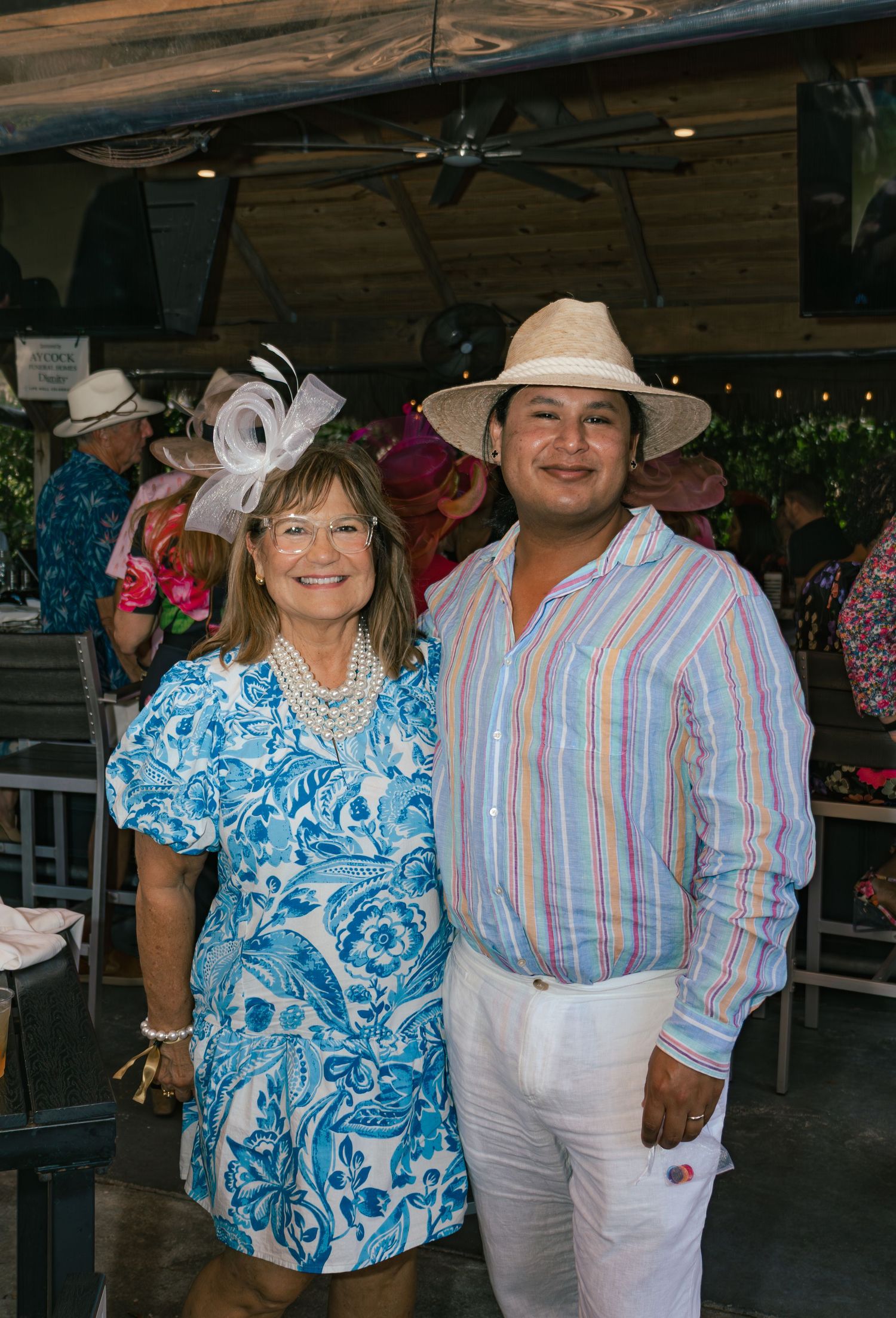 A woman in a blue floral dress and fascinator poses with a man in a striped shirt and panama hat.