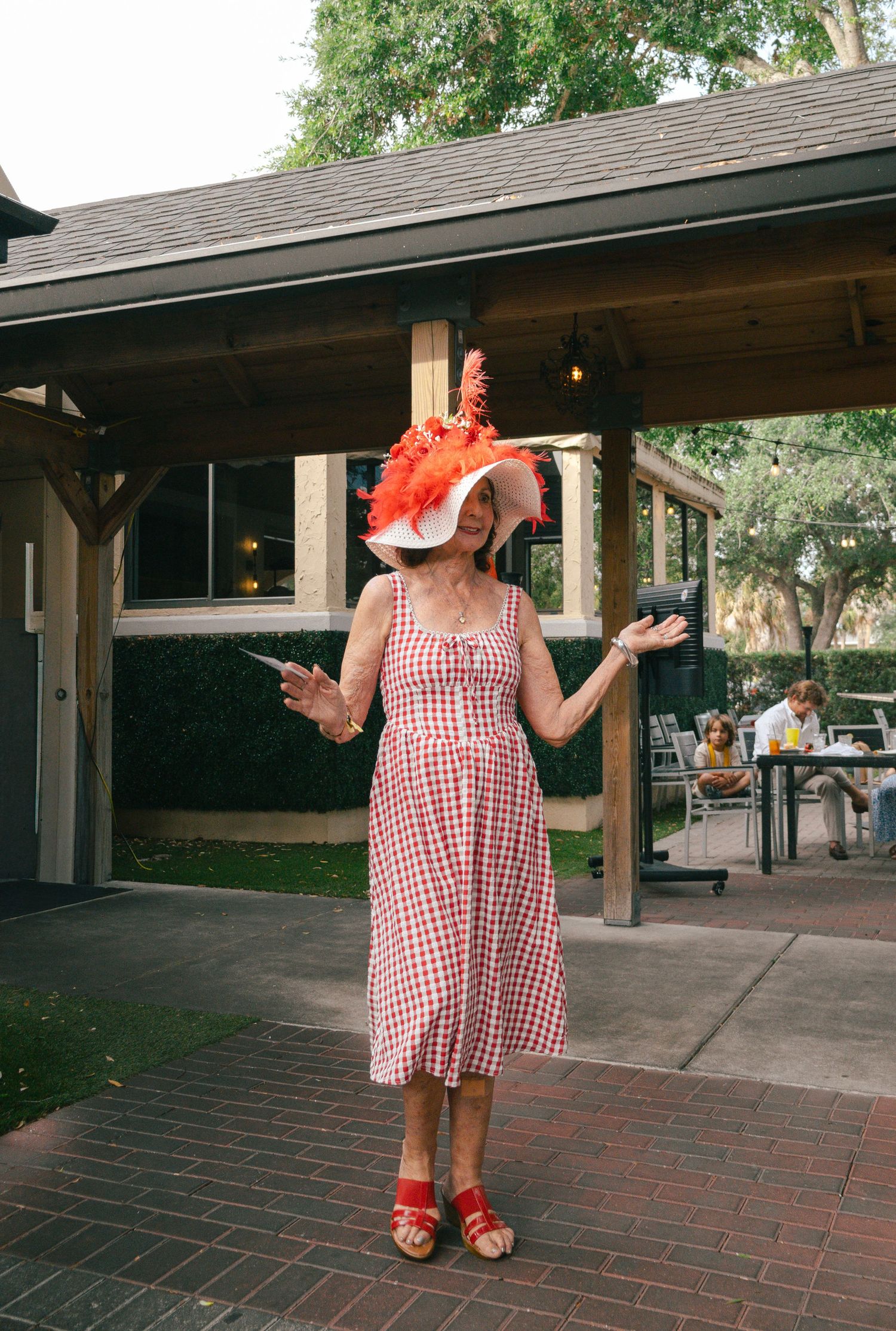 Woman in red gingham dress and large red feathered hat posing at an outdoor Kentucky Derby event.