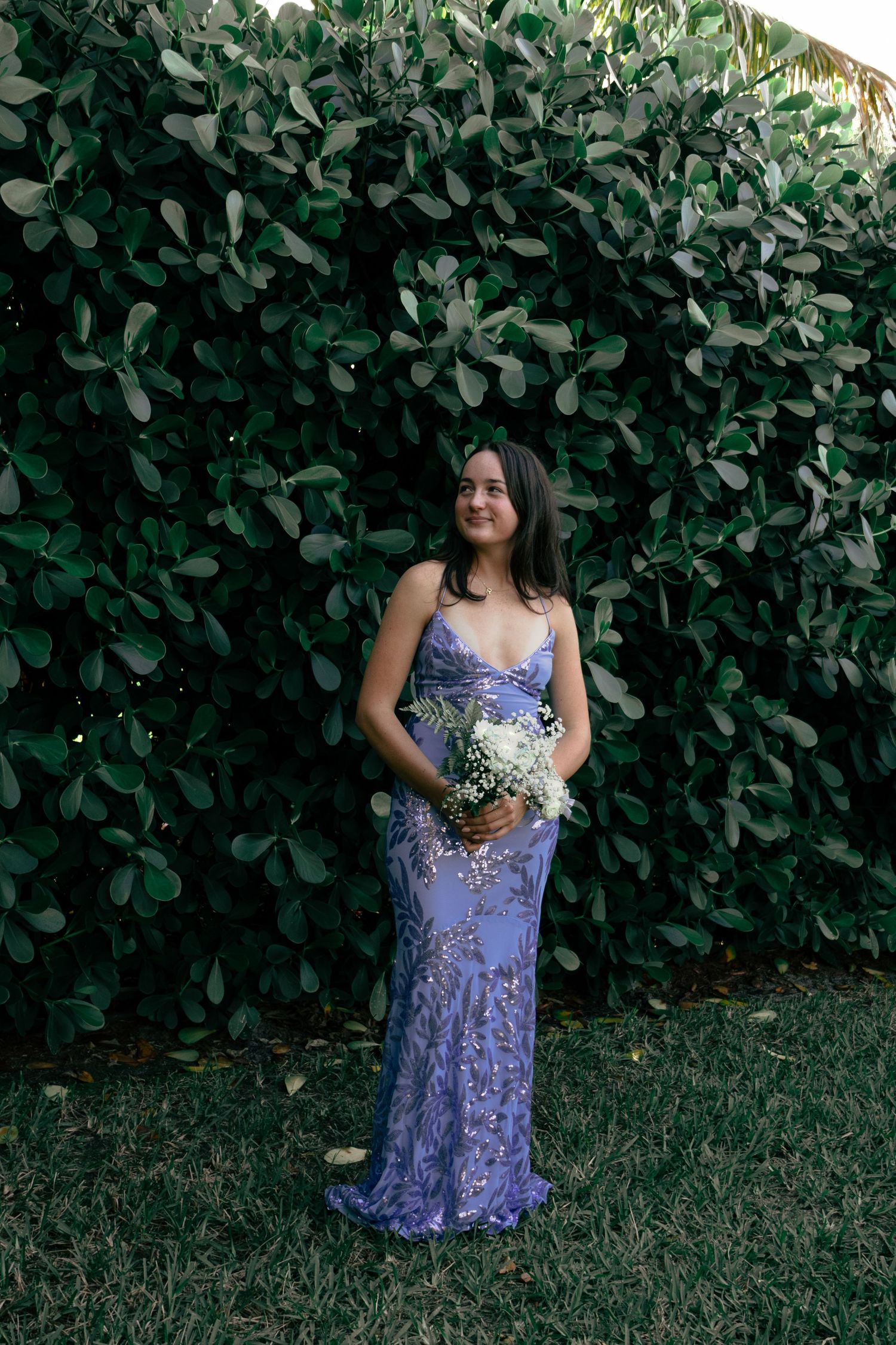 Young woman in a lavender sequined gown holding white flowers in front of a green hedge.