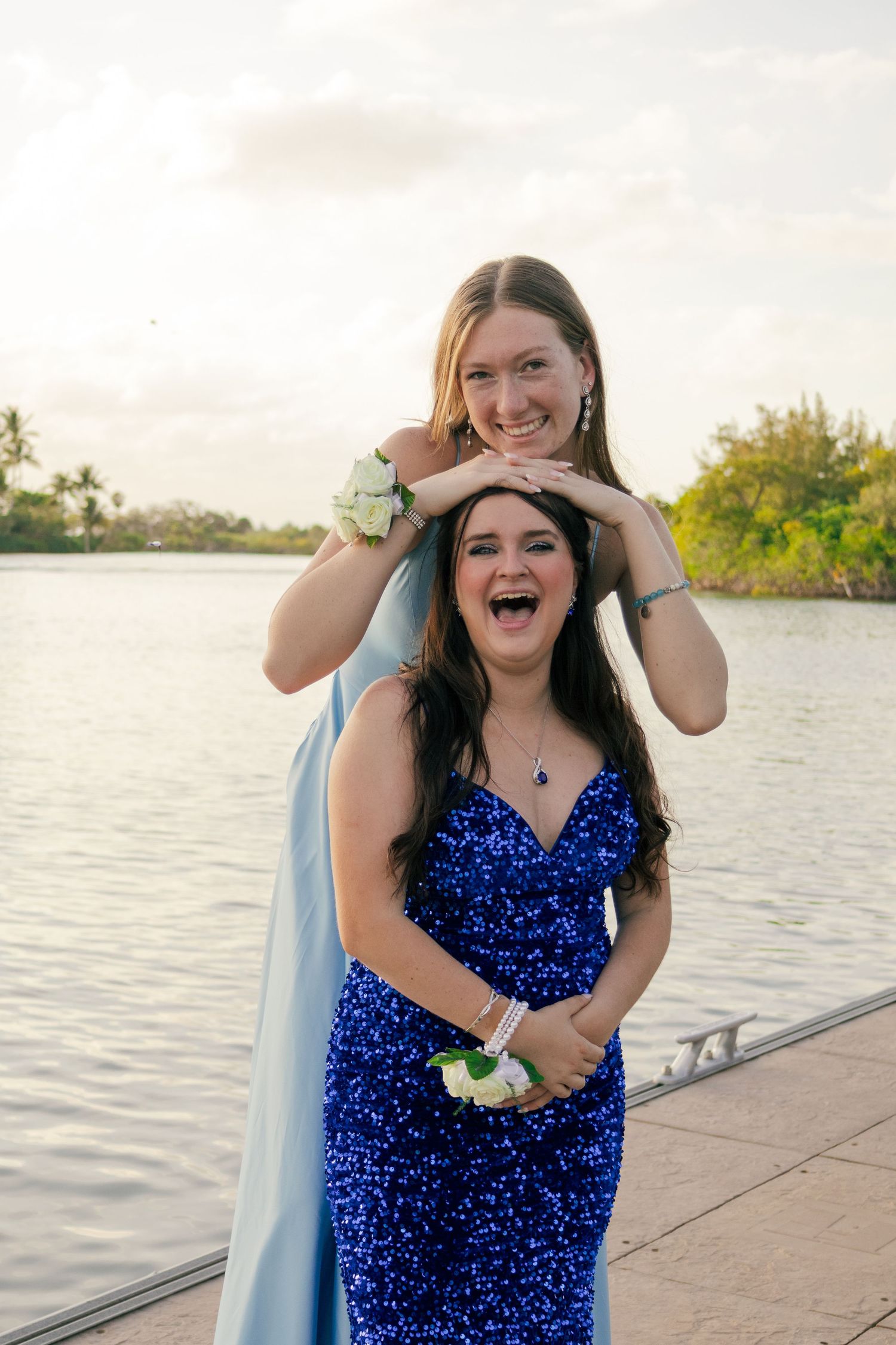 Two teenage girls in blue prom dresses laugh playfully together on a scenic waterfront dock.
