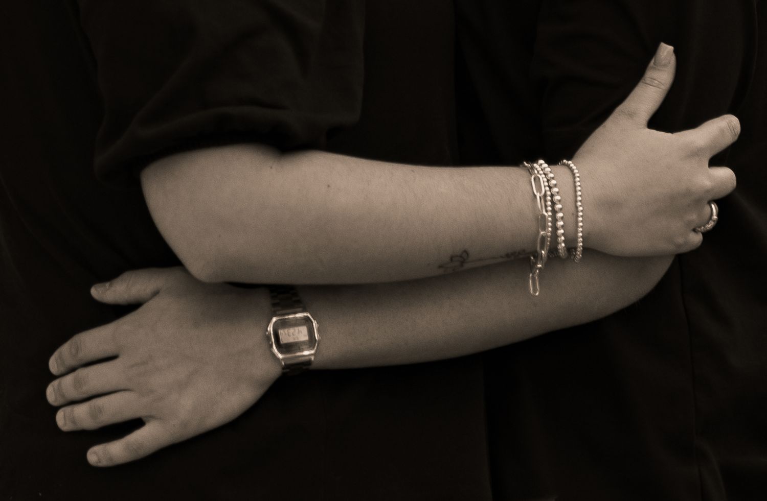 Close-up of intertwined arms showing bracelets, rings, and a digital watch against a dark background.