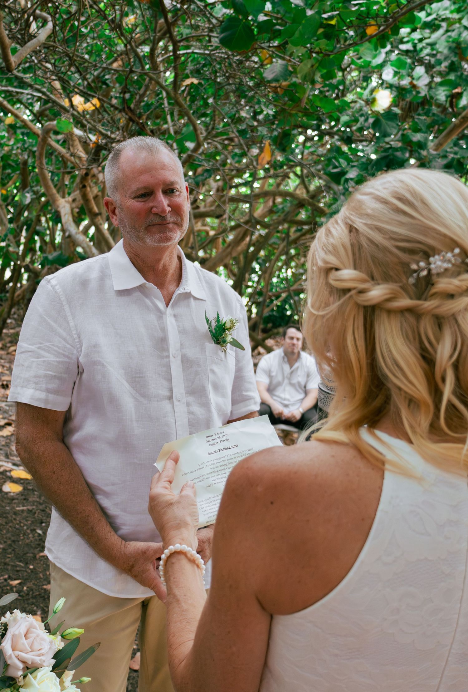 Groom smiling during outdoor wedding ceremony under lush green trees while bride reads vows.