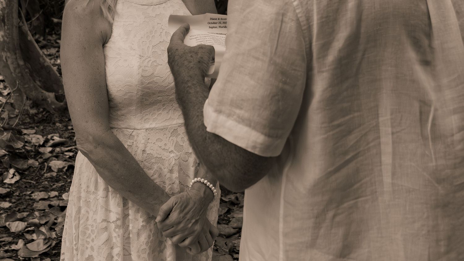 Bride and groom hold hands during wedding vows in sepia-toned photo.
