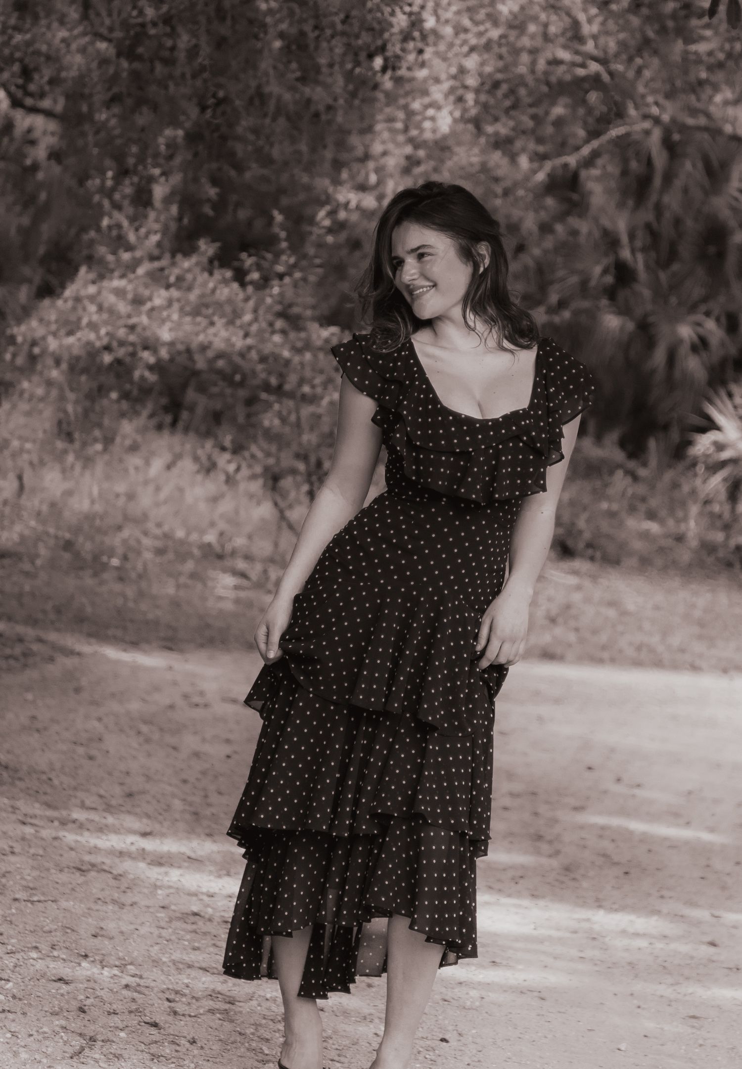 Black and white photo of young woman in polka dot dress smiling while walking on a dirt path.