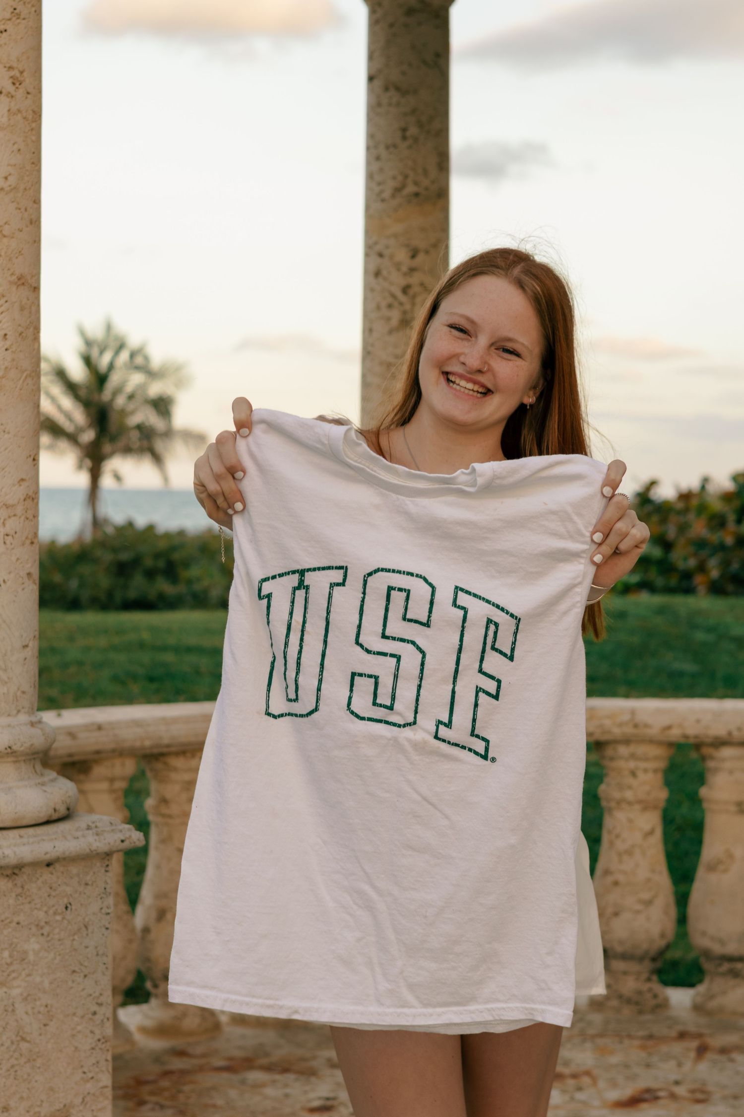 Smiling redhead girl holding white USF college t-shirt outdoors near ocean at sunset.