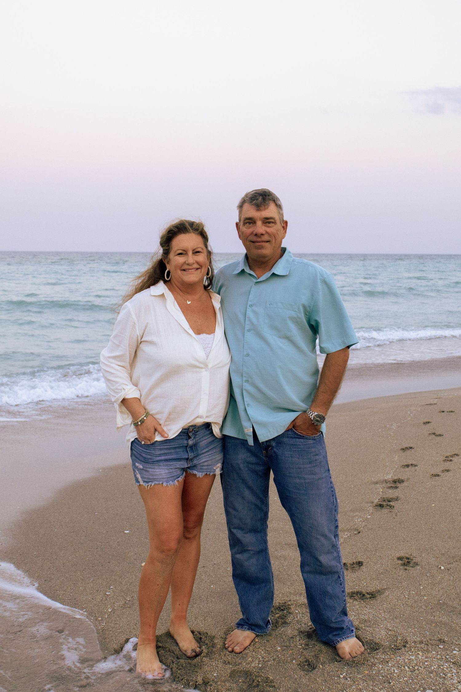Smiling couple standing together barefoot on sandy beach with calm ocean waves behind them.