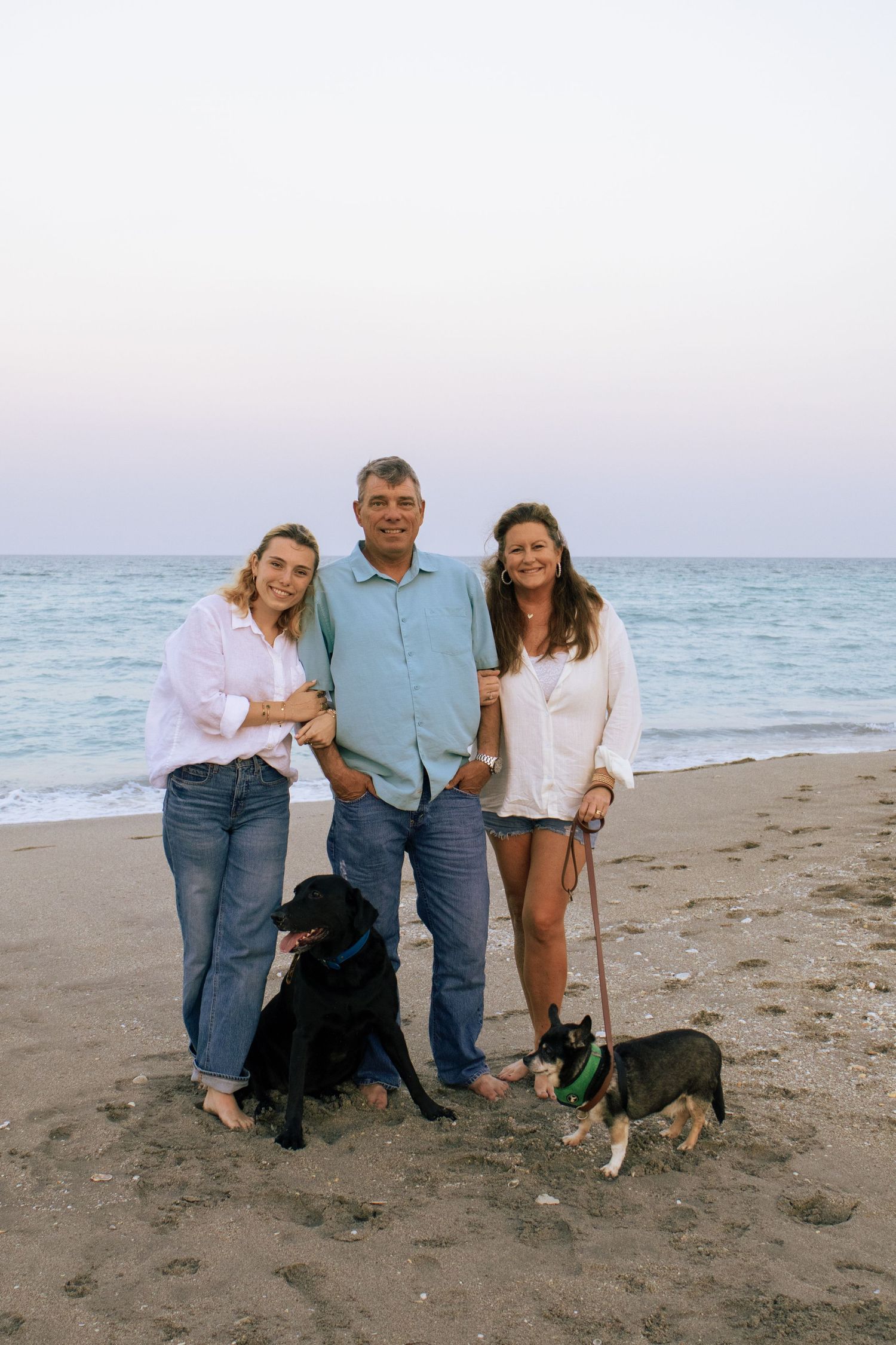 Family of three with two dogs posing on sandy beach at sunset with calm ocean waves in background.
