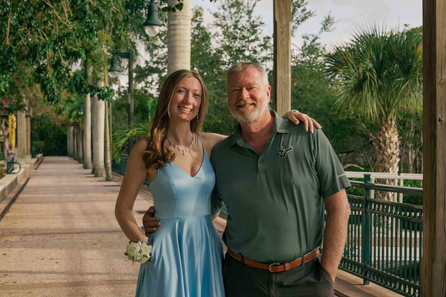 A young woman in a blue dress and an older man smile together under a columned outdoor walkway.