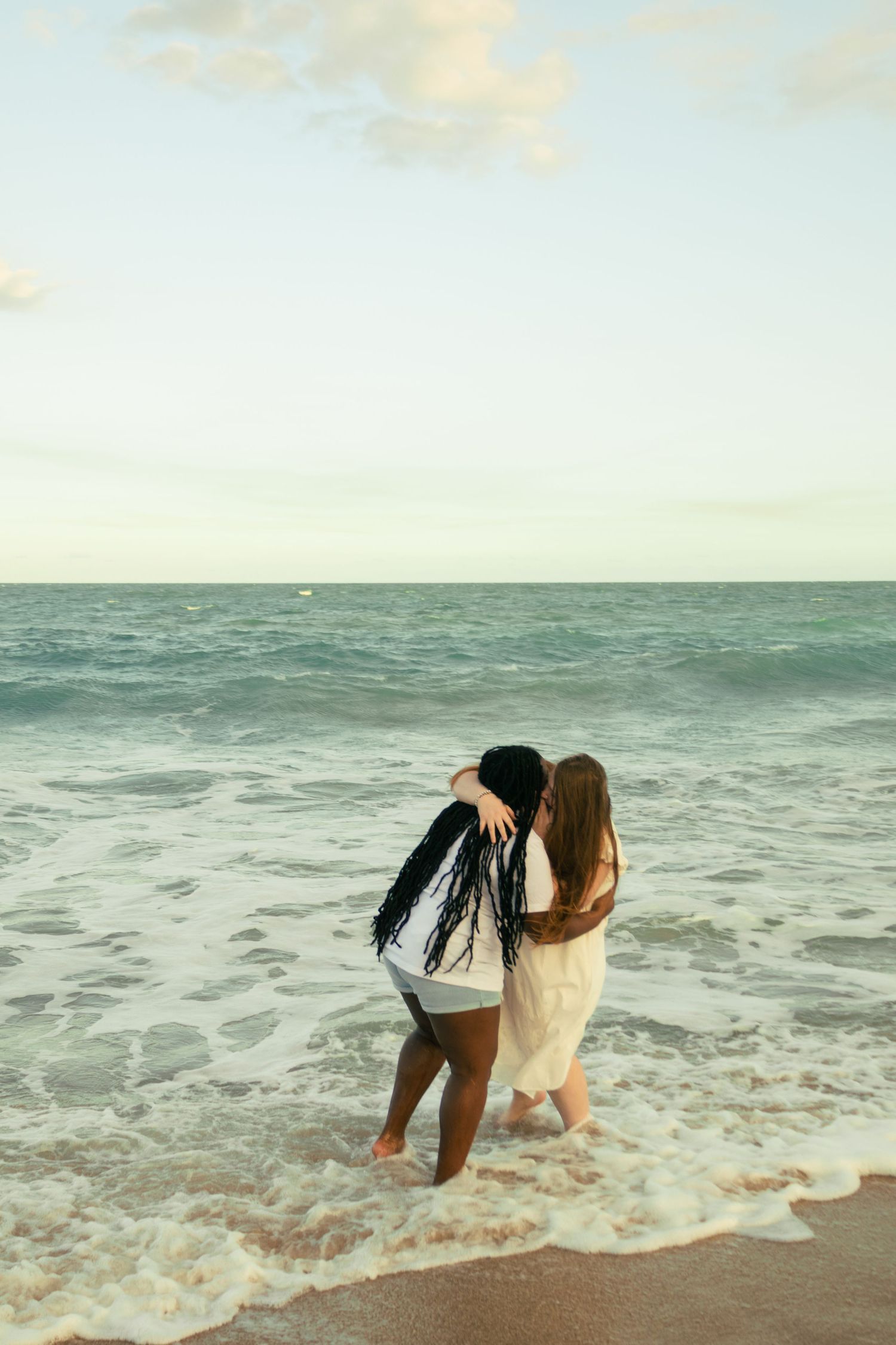 Two women hugging at the ocean's edge with waves around their feet.
