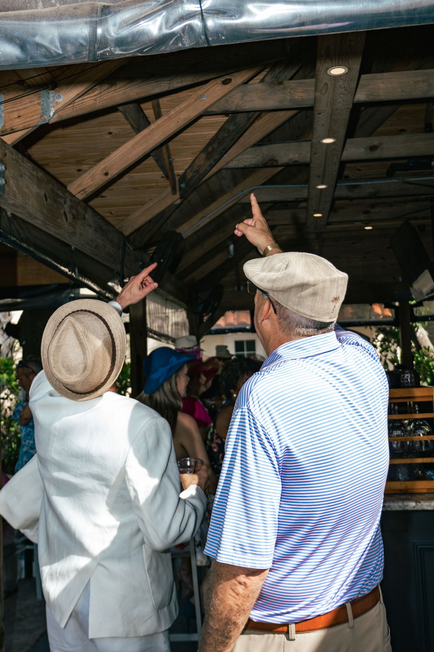 Two people in hats pointing at a rustic wooden outdoor bar area during a sunny event.