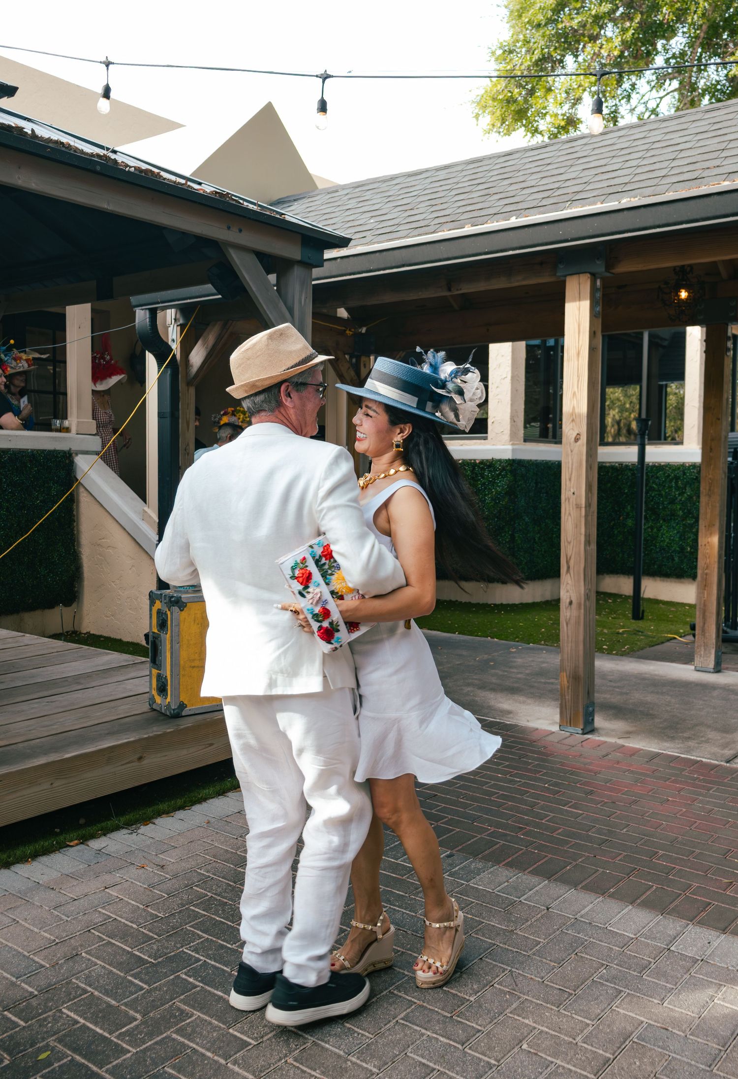 Couple in white Kentucky Derby attire, woman wearing blue fascinator hat, embrace on brick patio at outdoor event.